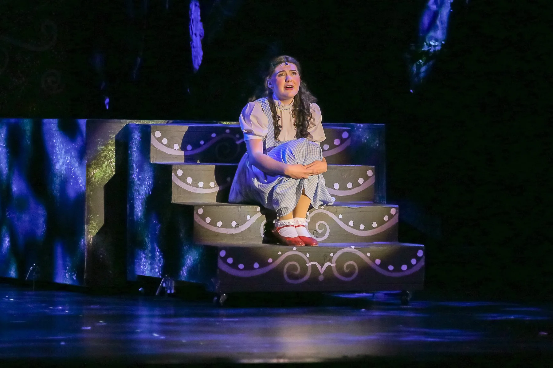 A young girl dressed in a costume with a plaid dress, white blouse, and red shoes is sitting on a decorated stage steps, holding her knees and looking upward with a worried or sad expression. The stage has black steps with purple and white swirl and 