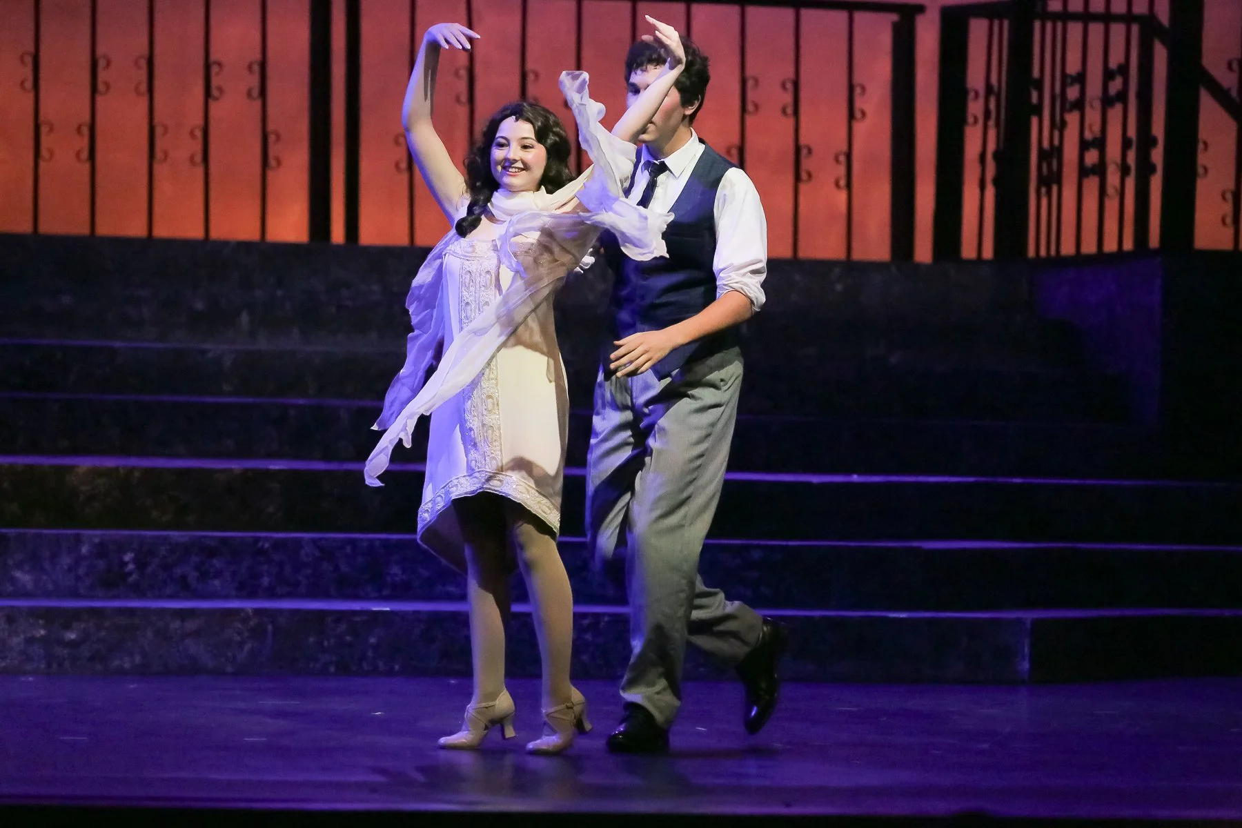 Two actors performing a ballet on stage, with a woman in a white dress and a man in a vest and trousers, against a backdrop of staircase and railing, illuminated by stage lights.