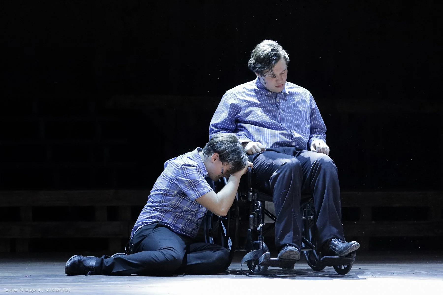 A person in a wheelchair with a serious expression, sitting on stage, with another person kneeling and holding their hand, creating an emotional moment in a performance.