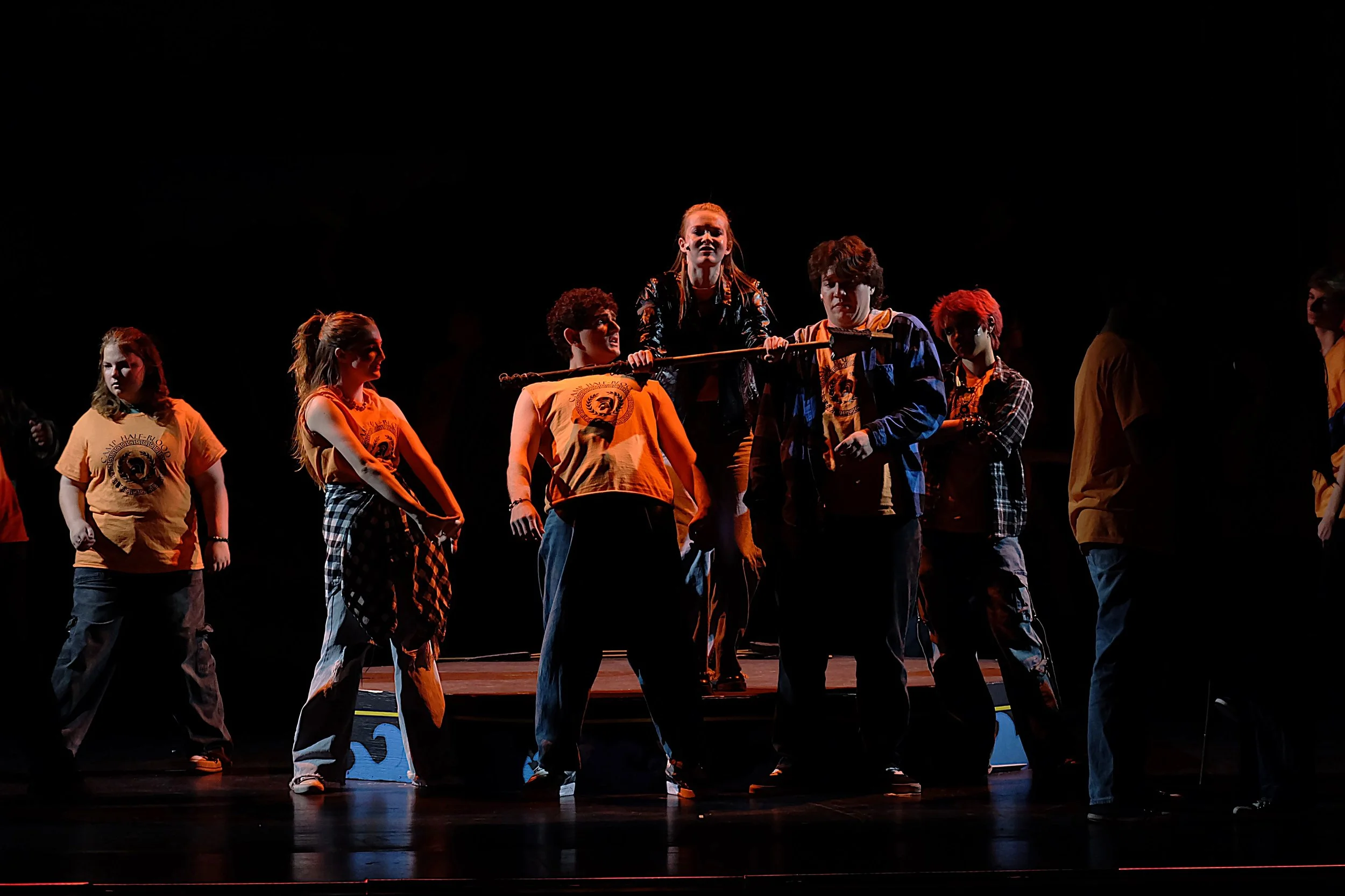 A group of young performers on stage, with one girl holding a staff, engaged in a theatrical performance with dark background and stage lighting.
