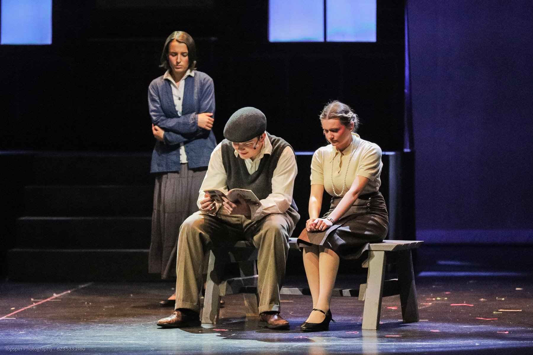 Three women on stage performing a play; two seated on a bench, one standing behind them, all appearing serious or contemplative.