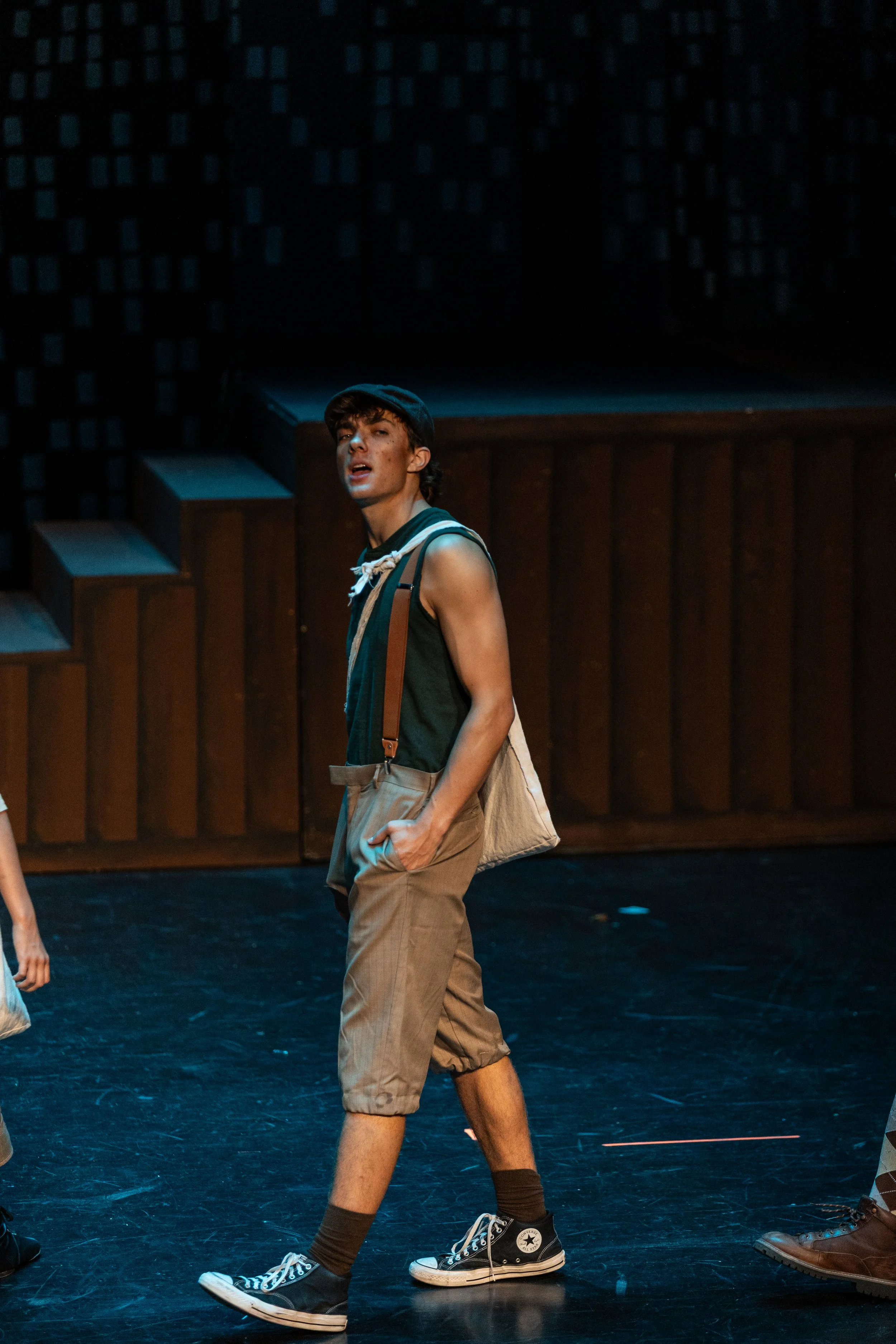 A young man with curly dark hair, wearing a dark cap, sleeveless shirt, tan shorts, black high-top Converse sneakers, and brown socks, standing on a stage with a wooden backdrop. He appears to be performing or speaking, with his mouth slightly open a
