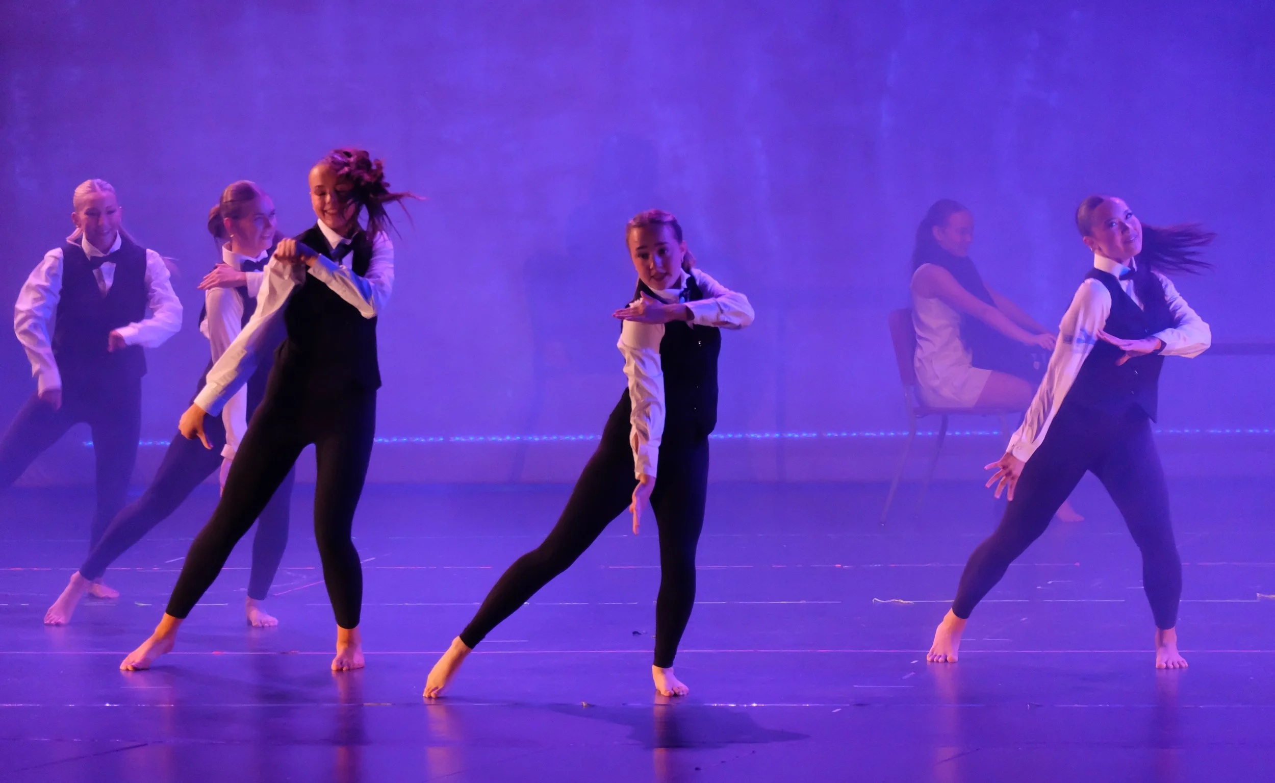 A group of six dancers performing on a stage with purple lighting, wearing black and white costumes with vests and pants, in a choreographed dance formation.