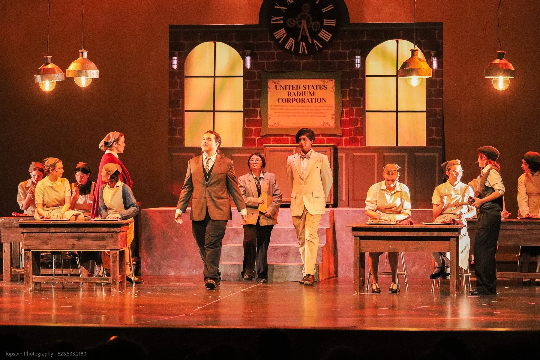 A theatrical stage with actors performing a historical scene. The set includes a backdrop of a brick wall, large windows, and a sign reading 'United States Radium Corporation.' Several actors are dressed in period costumes, and some are working at de