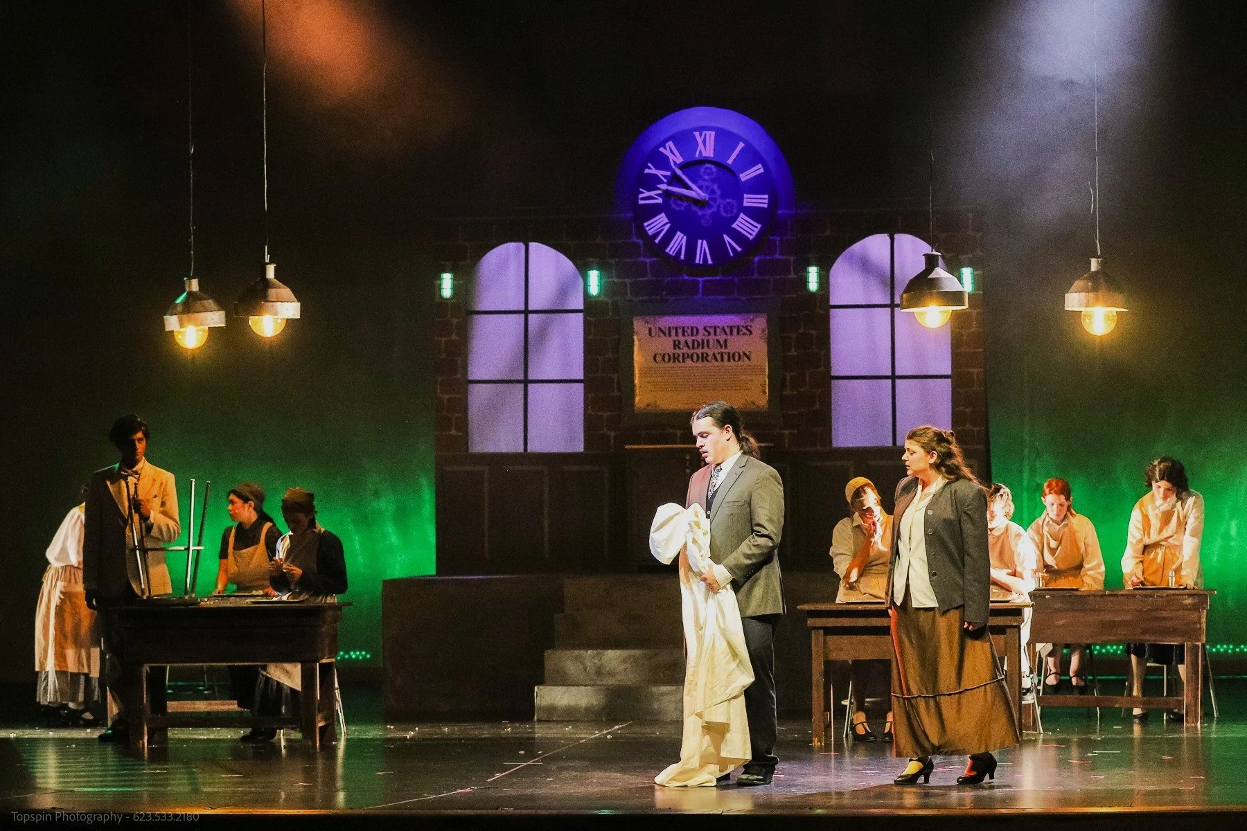 A theater stage scene depicting people in period costumes, some engaging in conversation and others working at tables, with a backdrop resembling an old factory or industrial building with a large clock reading 10:12 and a sign that says "United Stat