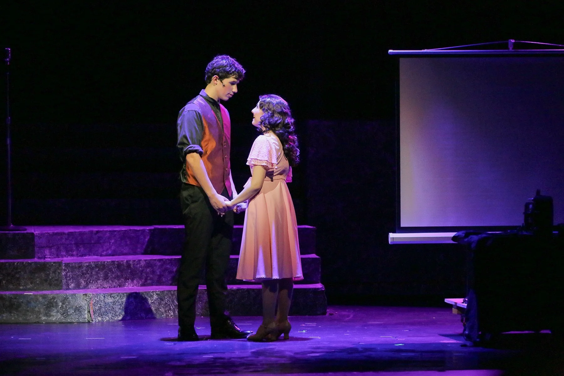 Two young actors, a boy and a girl, are on stage holding hands, looking into each other's eyes during a performance. The boy is wearing a black and red jacket with black pants, and the girl is dressed in a pink dress with curly hair. The stage has di