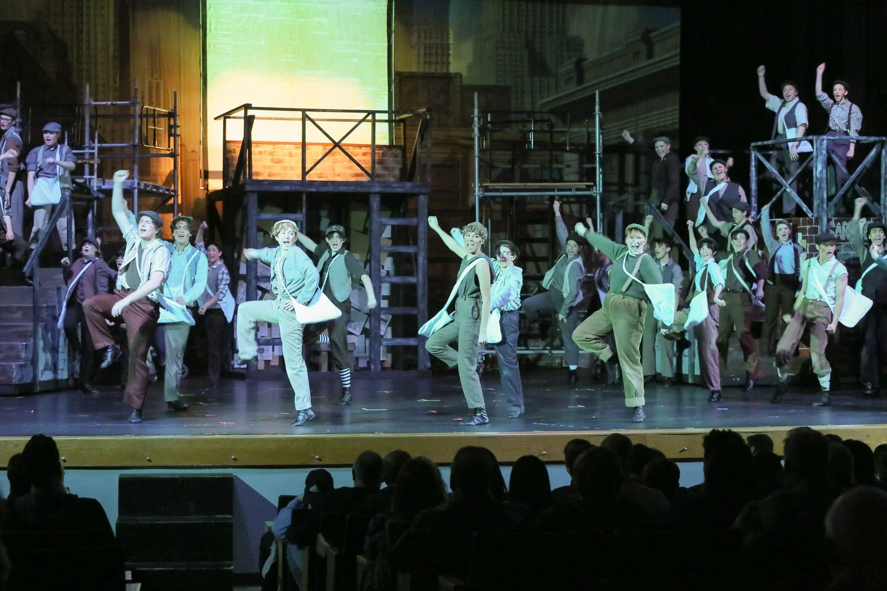 A theatrical stage with a cast of performers in period costumes. The background depicts an urban cityscape. Performers are mid-dance, smiling and energetic, with some raising their arms. The stage has multiple levels with scaffolding and set pieces.
