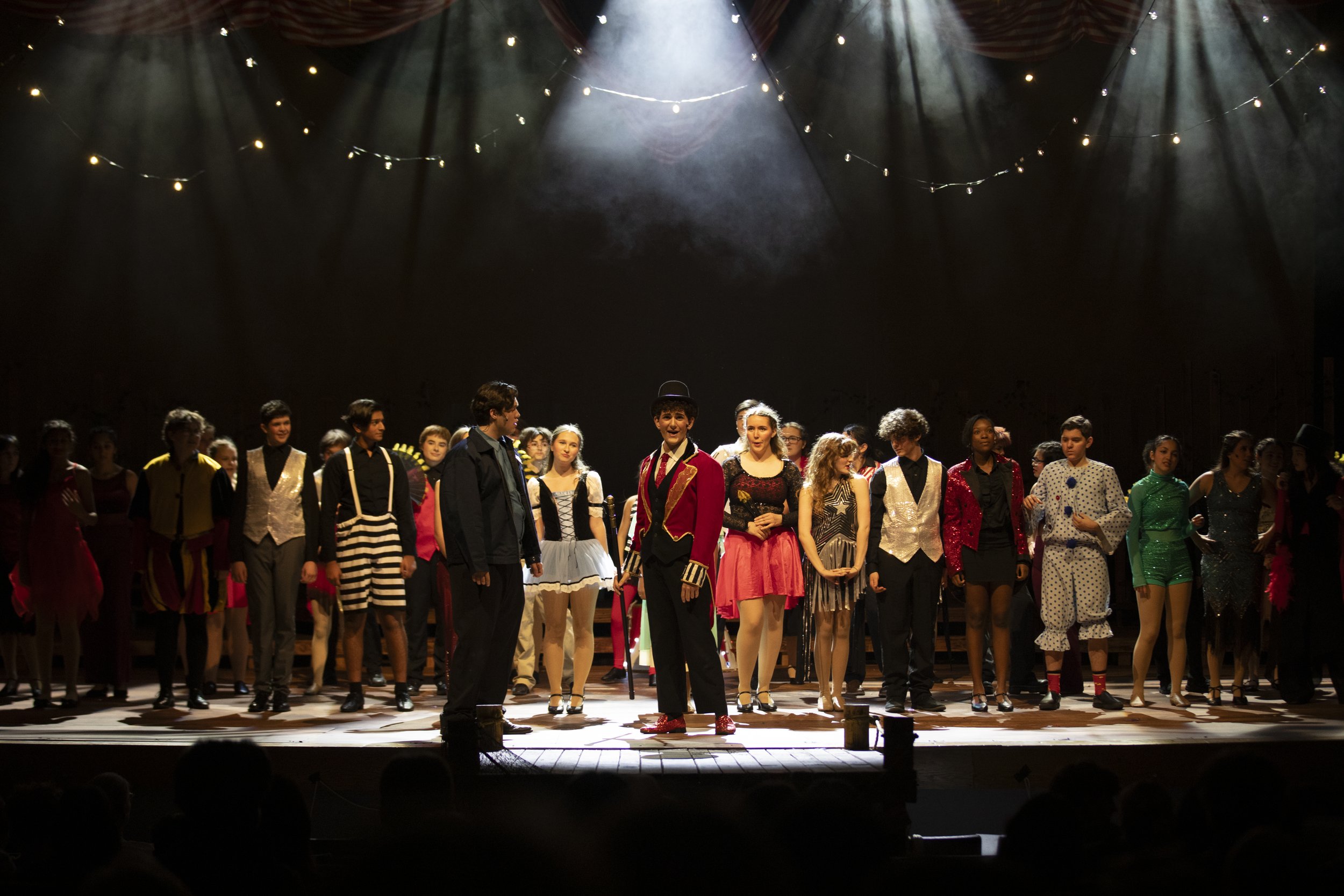 Group of children dressed in colorful costumes standing on a stage after a performance, with a dark backdrop and overhead stage lights.