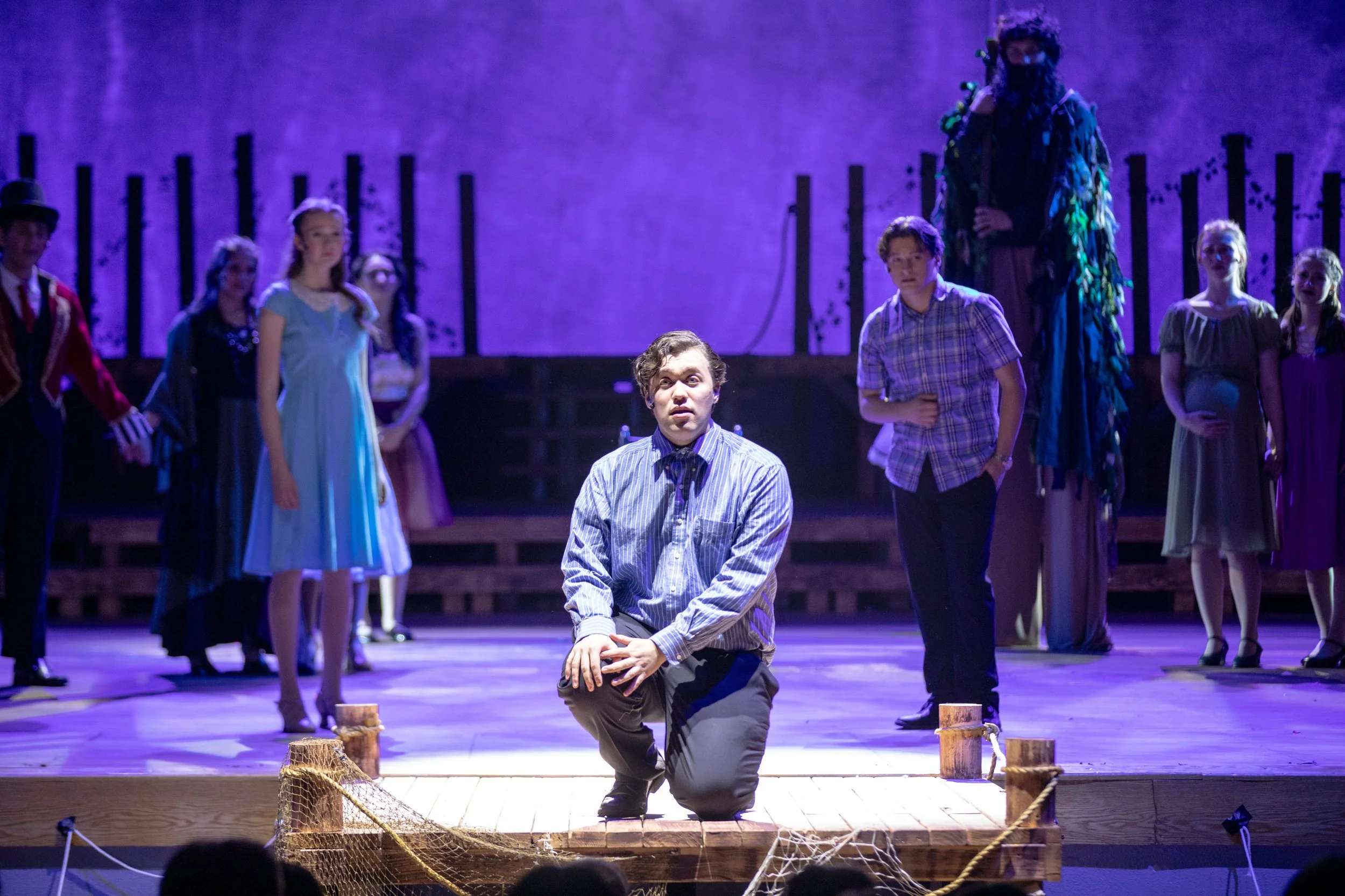 A theatrical stage performance featuring multiple actors in costume. One actor is kneeling in front, wearing a striped shirt, with other actors dressed in period costumes standing behind on stage with purple lighting. The scene seems dramatic and the
