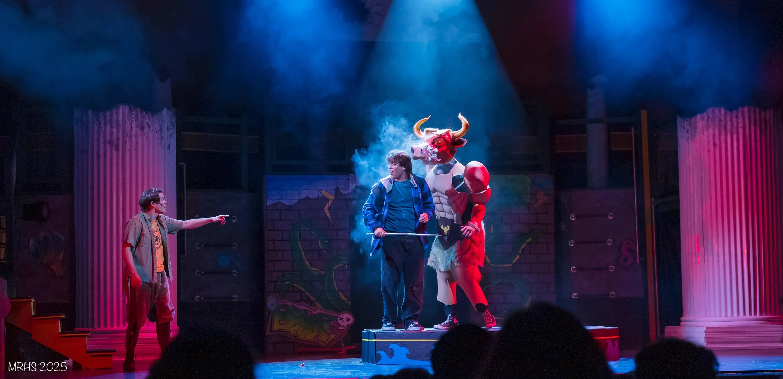 A theatrical stage scene with three characters: a person dressed as a dragon, a person with a walking stick, and another person pointing at the dragon, under colorful stage lighting.