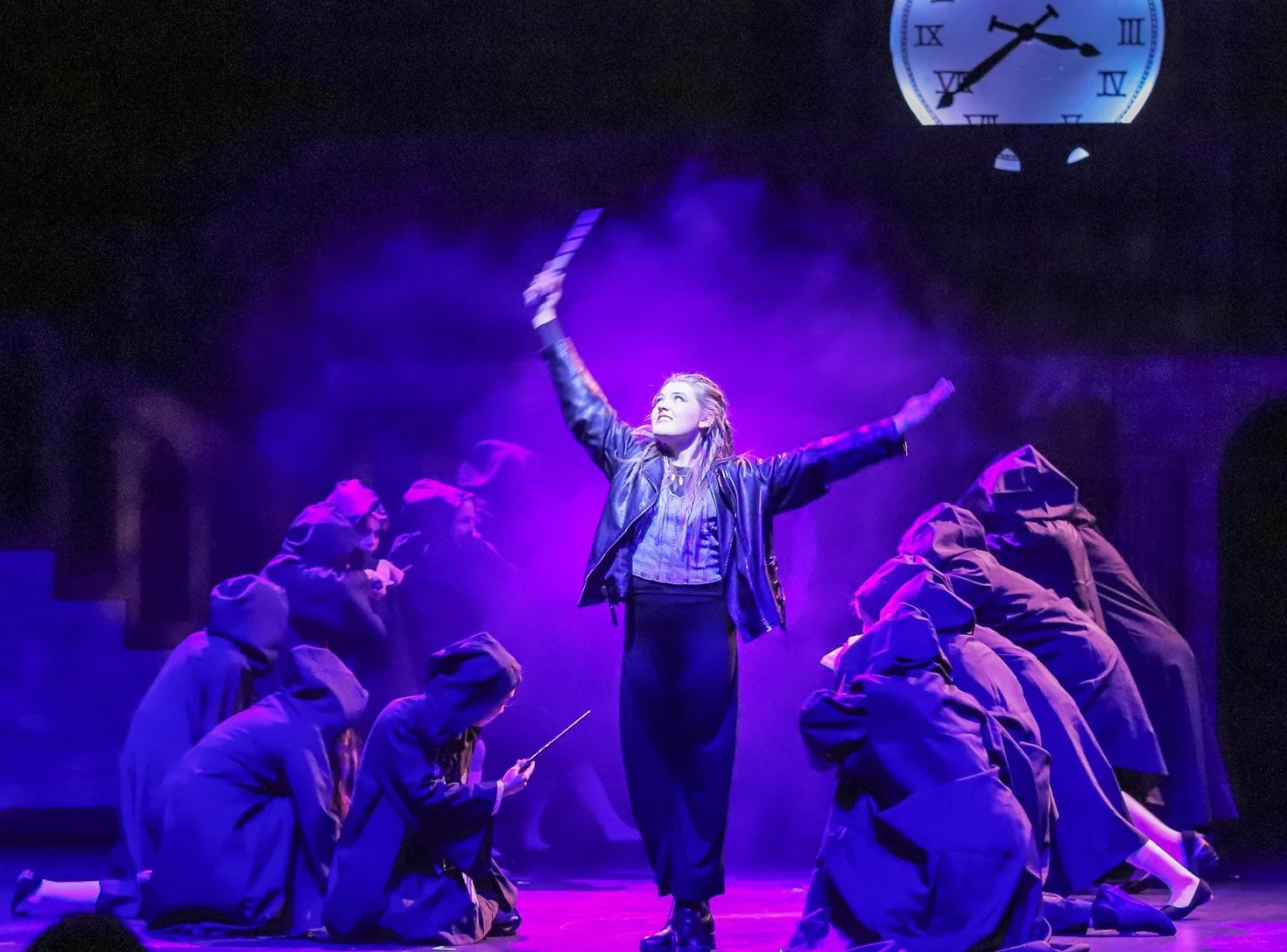 The image shows a theatrical stage scene with a woman in a leather jacket standing center, arms raised, surrounded by performers dressed in dark hooded cloaks. Purple lighting and fog create a dramatic atmosphere. A clock is visible in the background