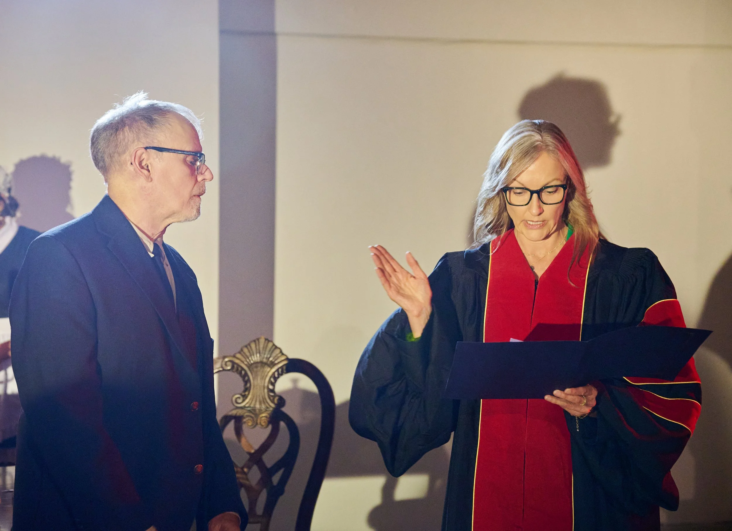 A man with glasses in a formal suit and a woman with glasses in a graduation gown are engaged in a conversation or interview, standing in a room with a neutral background and ornate decor.