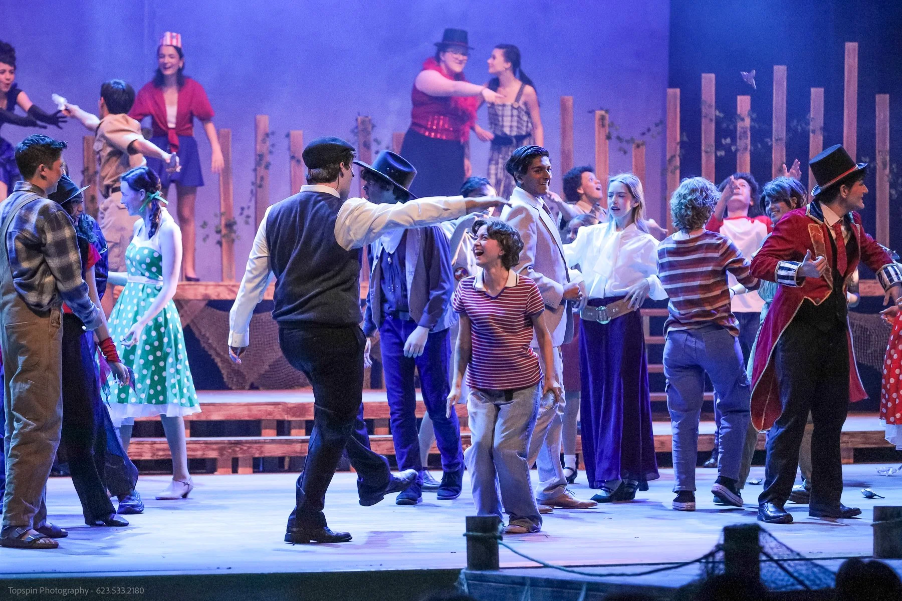 The image shows a cast performing on stage in a theatrical production, with actors dressed in colorful costumes, some in period clothing, engaging in dance and dialogue. The background features wooden fencing and stage props, with a purple hue lighti