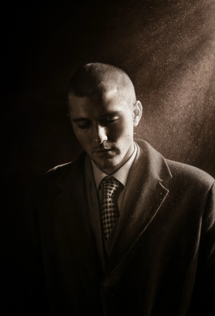 Robert Junqueira wearing a suit and tie, with short hair, has a contemplative expression as dust particles or powder are seen floating around in the air, illuminated by a light source behind him.