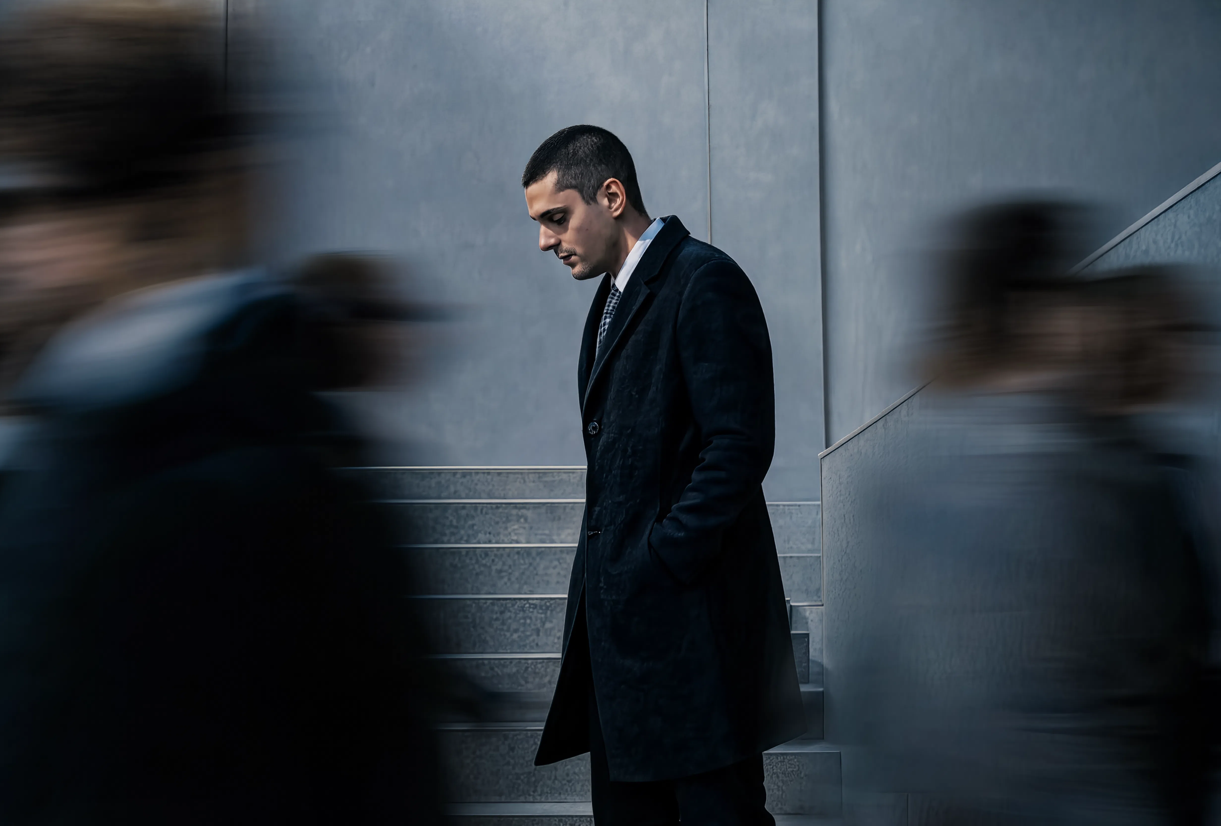 Robert Junqueira in a dark suit standing against a gray wall, looking down with a serious expression. Blurred figures pass by on either side.
