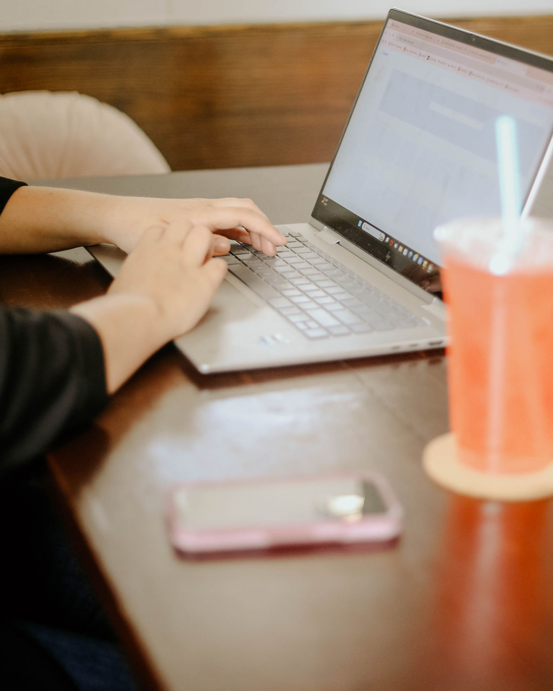 Person typing on a silver laptop with a pink smartphone and a pink drink with a straw on a wooden table.