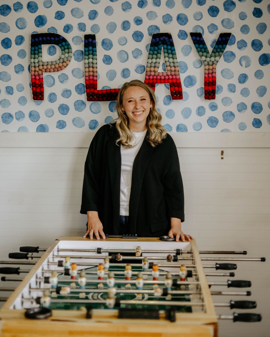 A smiling woman standing behind a foosball table in a room with a wall decorated with the word 'PLAY' in colorful bubble-like letters and blue polka dots.