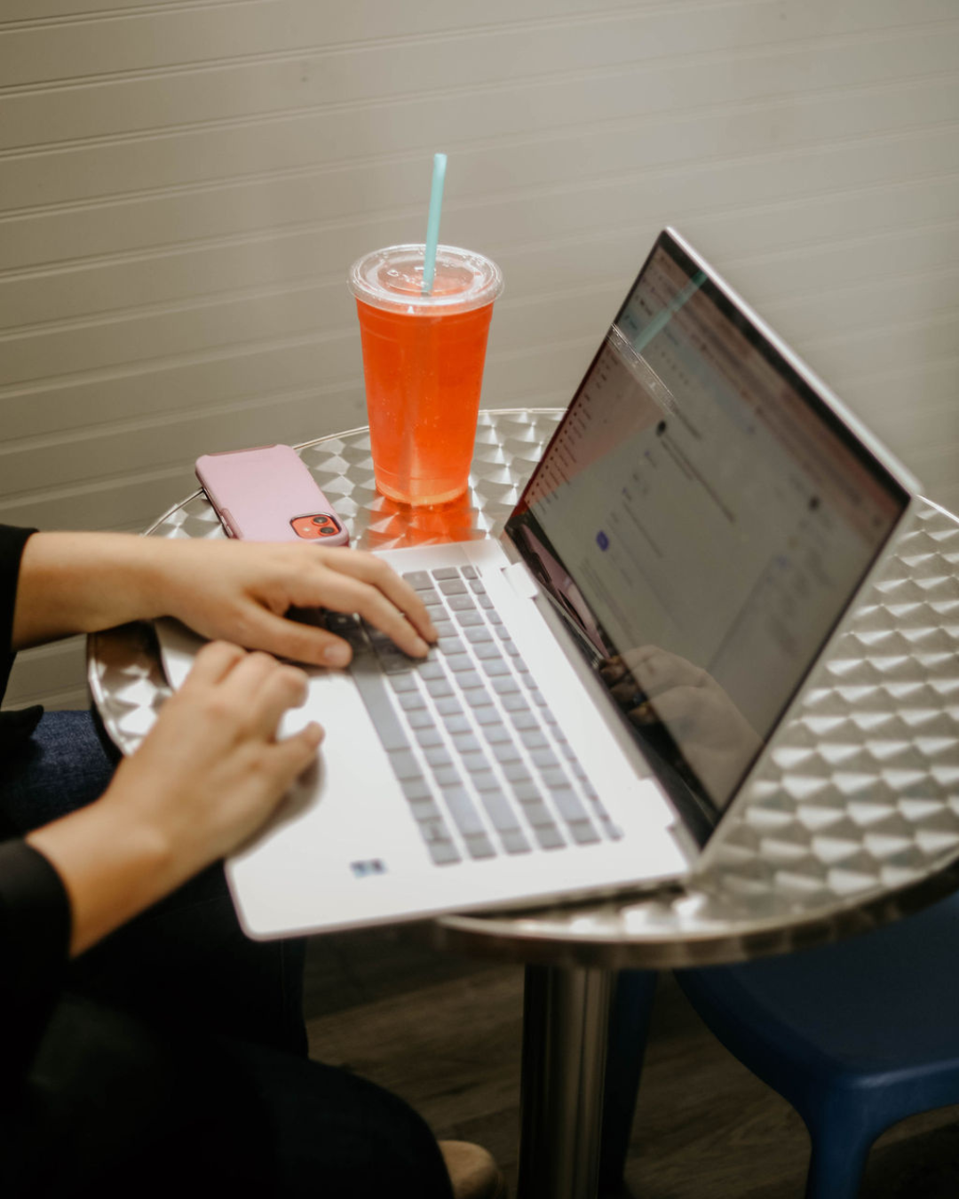 Close-up of a person typing on a silver laptop with a pink phone and a large cold drink with a straw on a small round metallic table.