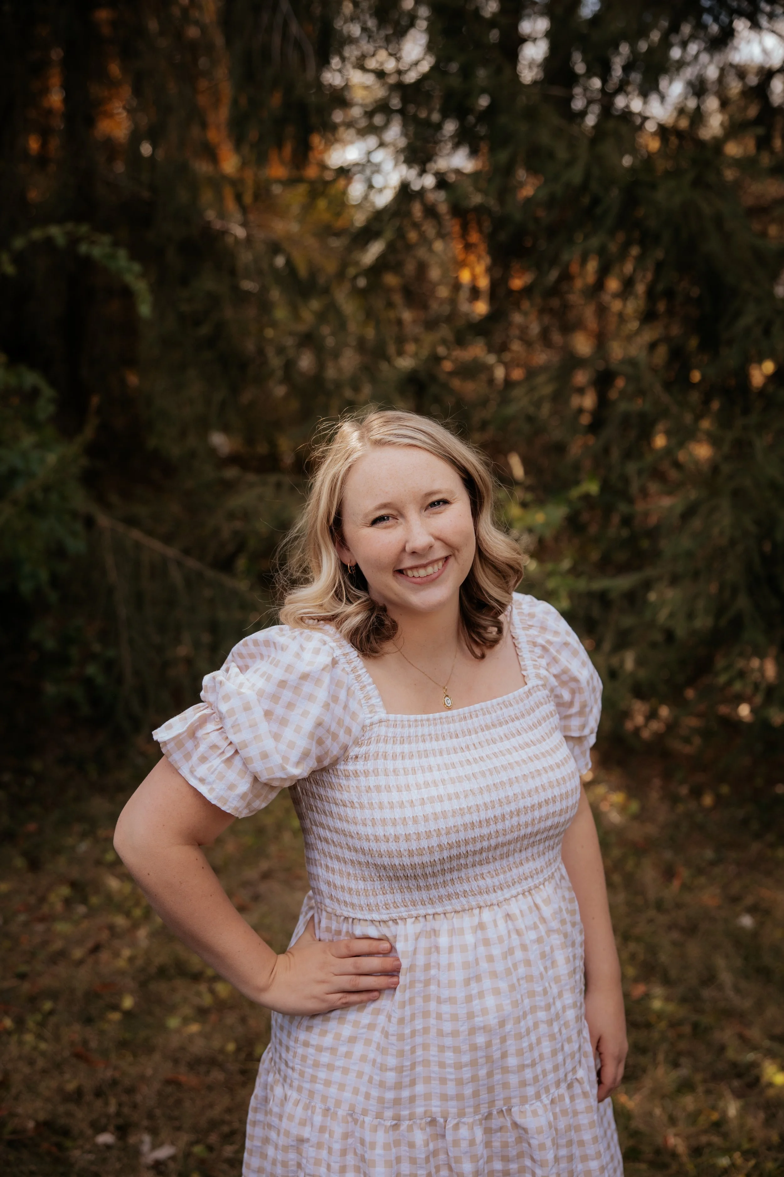 A young woman with blonde, wavy hair, wearing a light-colored checkered dress with puffed sleeves, smiling outdoors in front of a background of trees.
