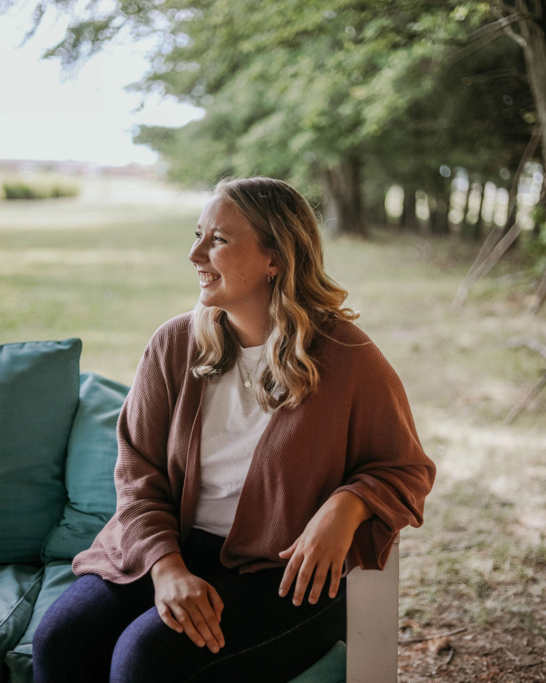 A woman with wavy blonde hair, smiling and sitting outdoors on a porch or patio with trees and grass in the background.