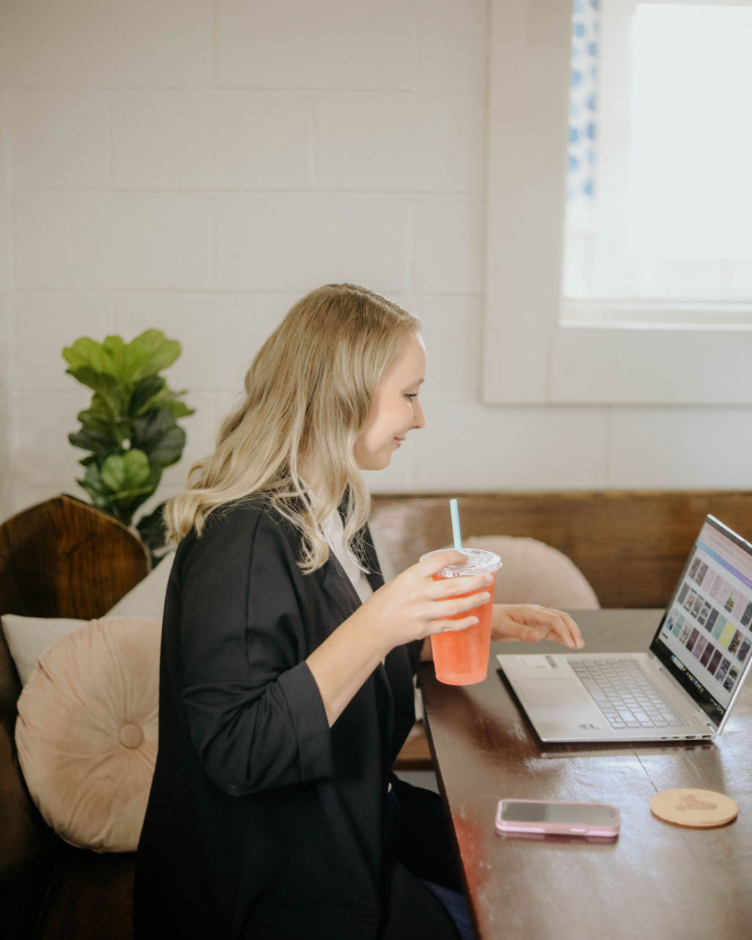A woman with blonde hair working on a laptop at a wooden table, holding a red drink with a straw, with a smartphone and a coaster nearby, in a cozy cafe or workspace.