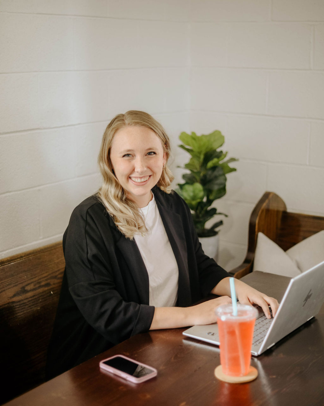 Smiling woman working on a laptop at a wooden table with a pink phone and a cup with a straw, in a room with white brick walls and a green plant in the background.