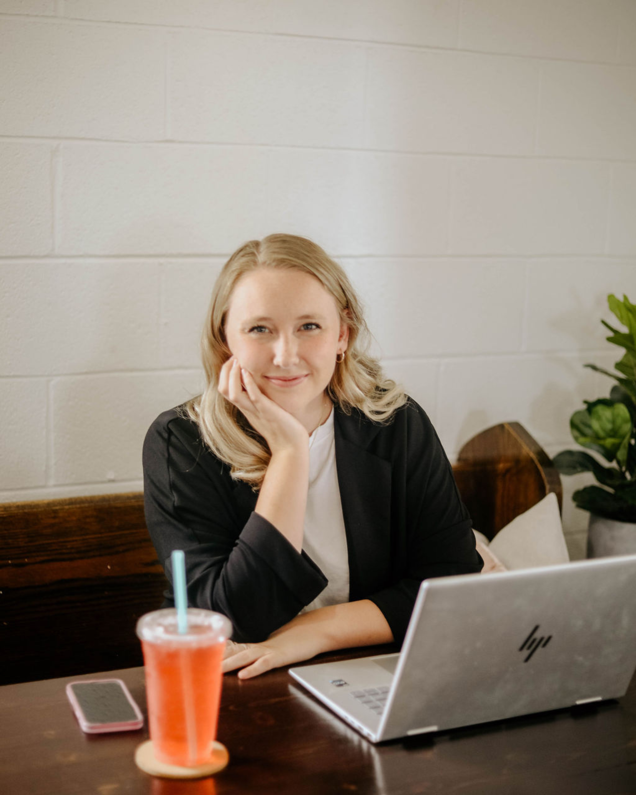 A woman sitting at a wooden table with a gray HP laptop, a pink smartphone, and a red drink with a straw.
