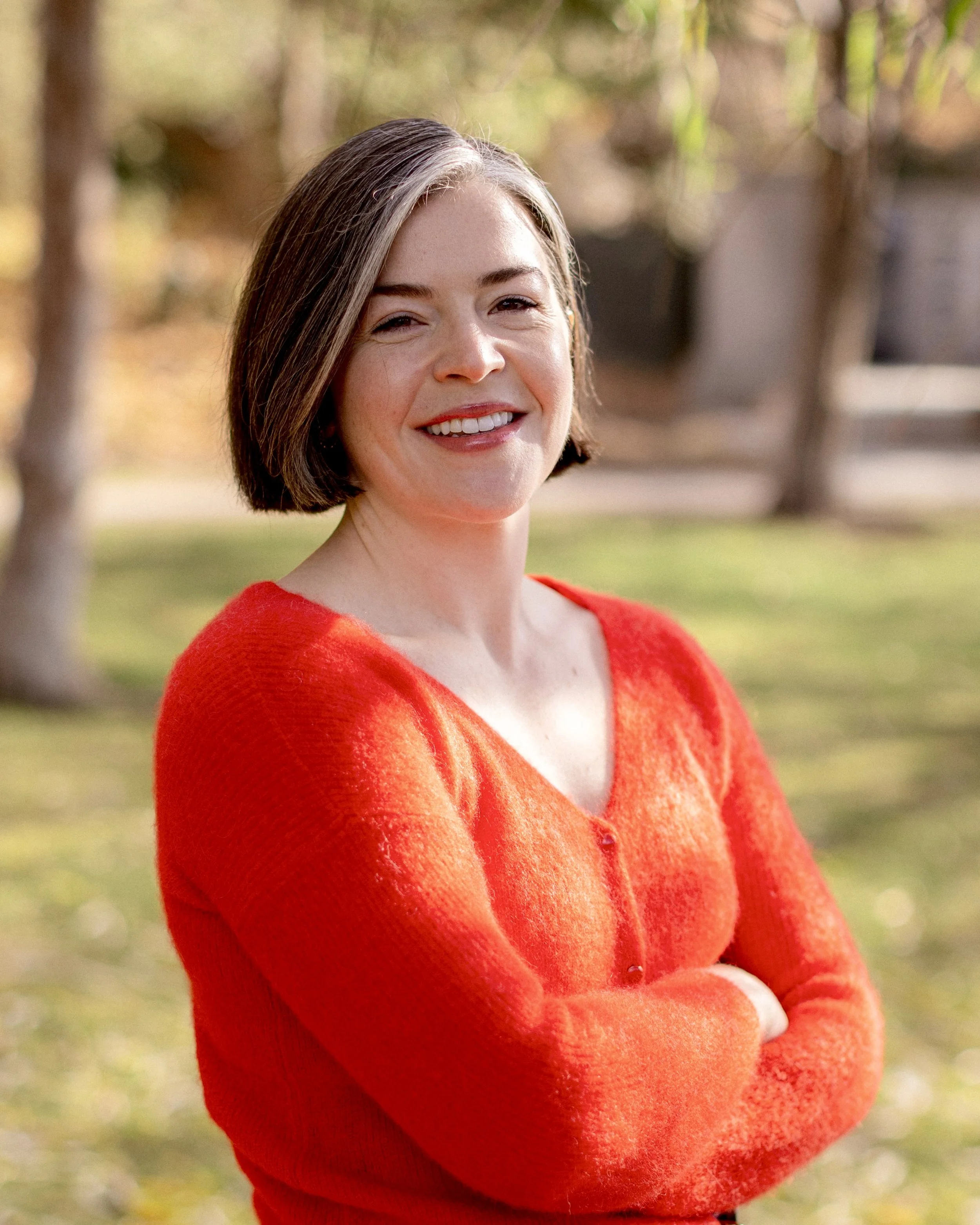 A woman with short brown hair, wearing a red sweater, smiling outdoors in a park or garden setting.