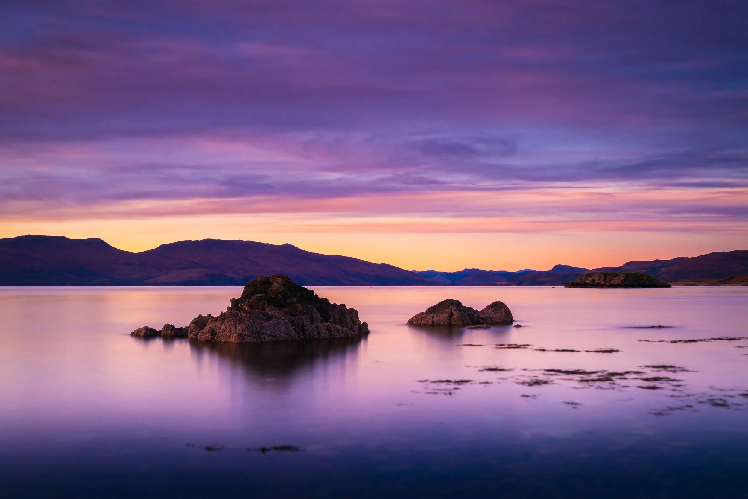Knoydart Peninsula, Scotland. Looking north towards the Kyle Of Lochalsh at sunset with a colorful sky displaying shades of purple, pink, and orange, surrounded by distant mountains and rocks in the foreground.