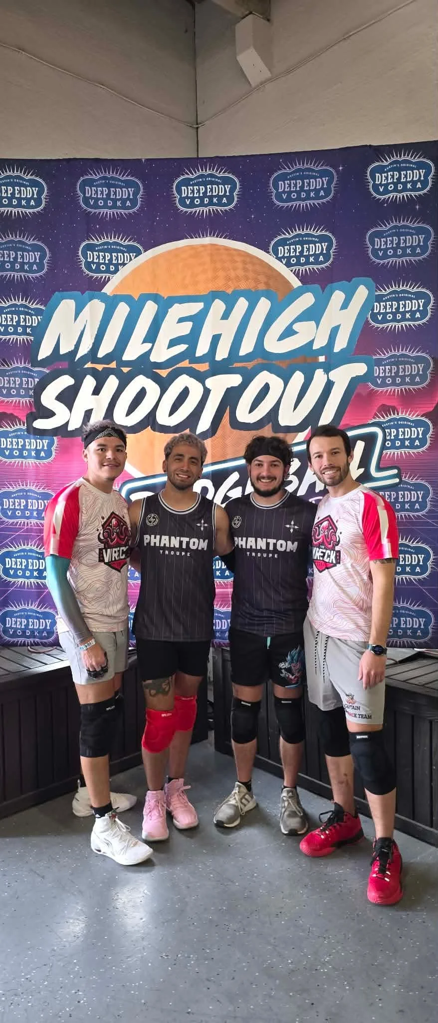 Four men standing in front of a colorful banner at the MileHigh Shootout event. The banner features the Deep Eddy Vodka logo and the event name in large letters, with a sunset graphic in the background. The men are dressed in a mix of casual and sportswear, some wearing jerseys that say "Phantom" and others in team themed attire.