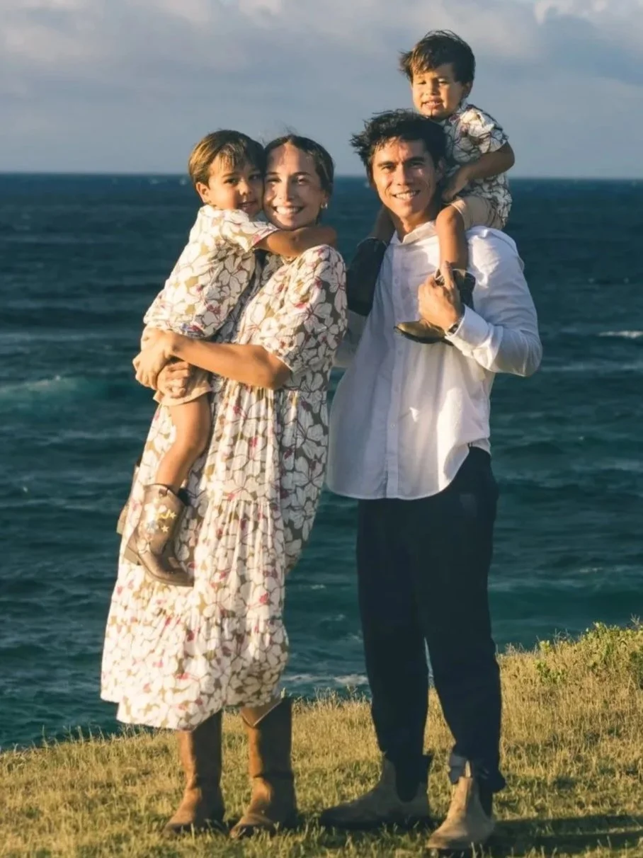Family of four outdoors by the ocean, with two children on the parents' shoulders, smiling and enjoying the sunset.