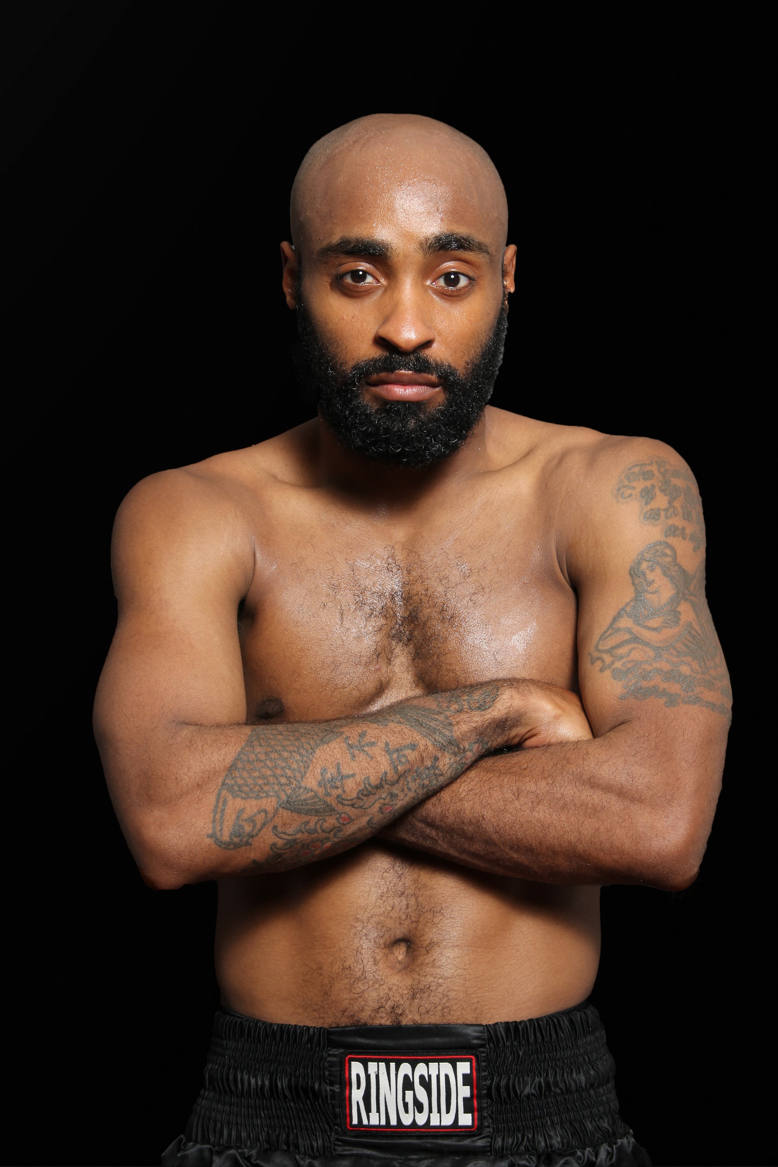 A shirtless man with a beard, tattoos on his arms and chest, crossing his arms in front of his chest, against a black background.