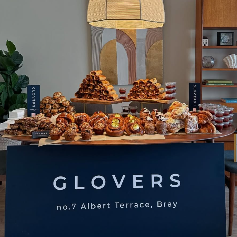 A table with an assortment of baked goods including croissants, muffins, cinnamon rolls, and small cups of jam, with a large sign in front that reads "GLOVERS no.7 Albert Terrace, Bray." Online ordering signs labeled "GLOVERS" are on each side of the table, and a large lamp and abstract art hang on the wall behind the table.