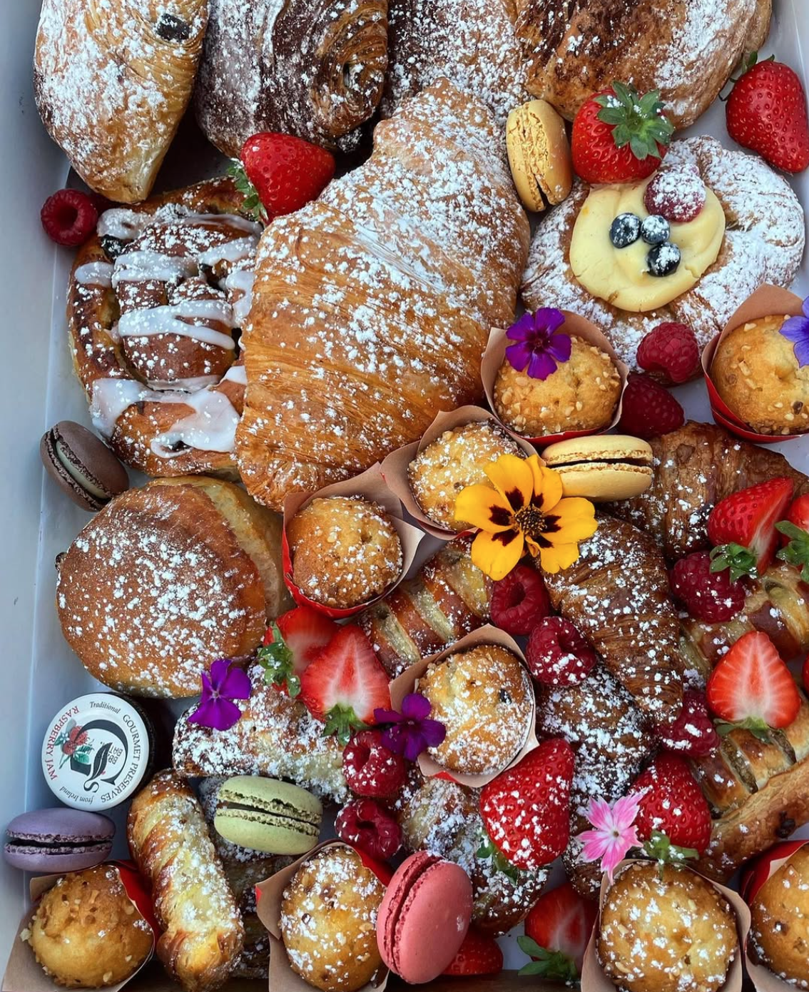 An assortment of baked goods including croissants, muffins, cookies, and cupcakes, garnished with fresh strawberries, raspberries, blueberries, flowers, and macarons.