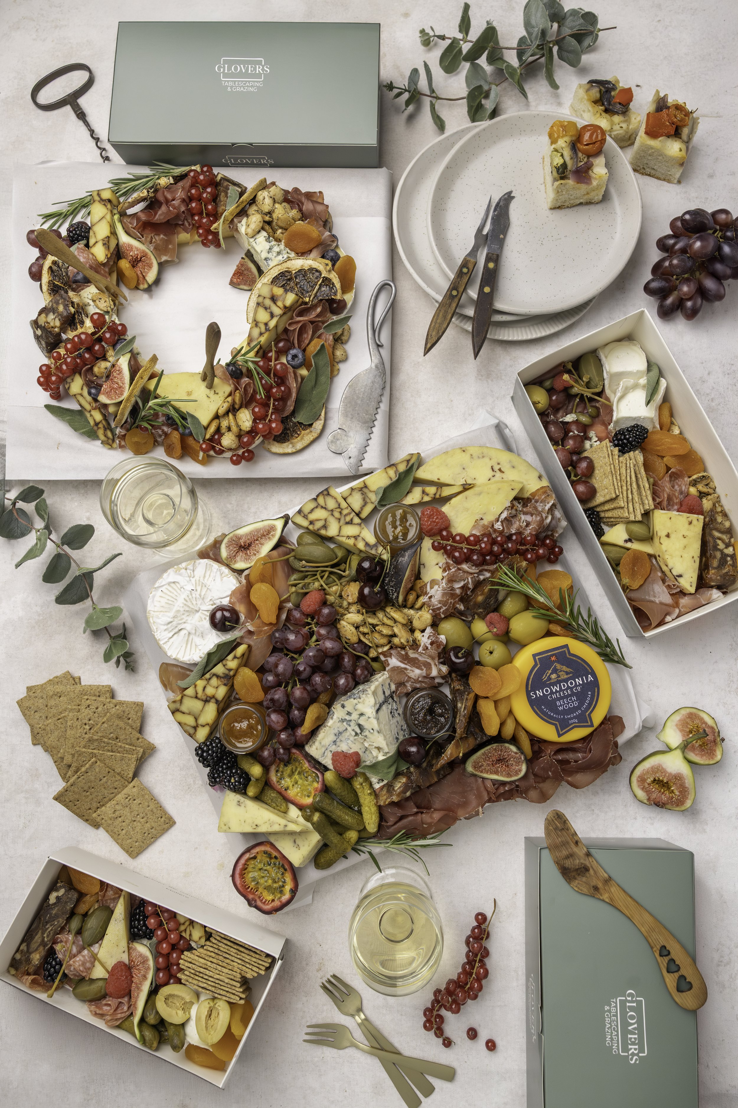 Charcuterie boards with cheeses, grapes, figs, crackers, and charcuterie meats, topped with herbs and dried fruits. Small plates with appetizers and glasses of white wine are also on the table, set for a gathering or celebration.