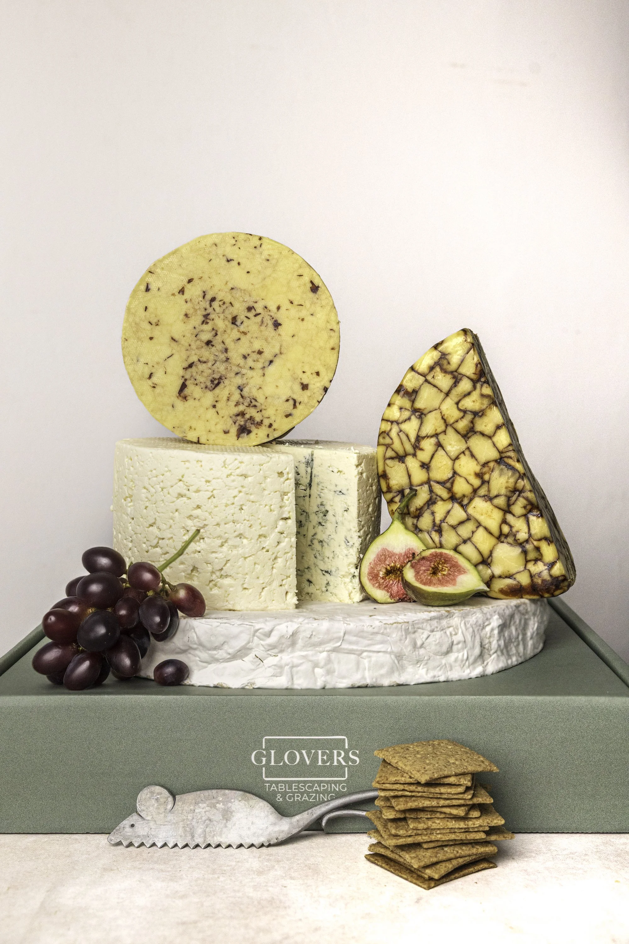 A cheese platter with grapes, figs, and slices of cheese, displayed on a green box labeled GLOVERS TABLESCAPING & GRAZING, accompanied by a small stack of crackers and a cheese knife.