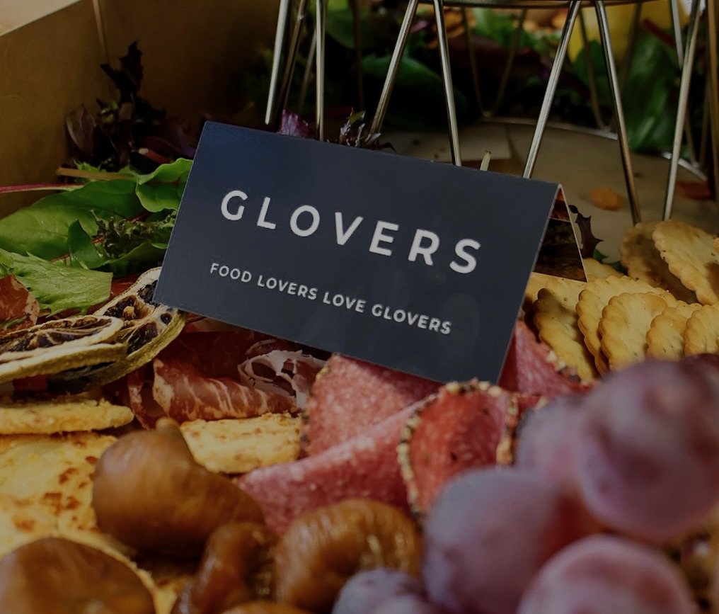 Close-up of a charcuterie board with cheese, cured meats, grapes, and nuts, with a sign that reads "GLOVERS FOOD LOVERS LOVE GLOVERS."