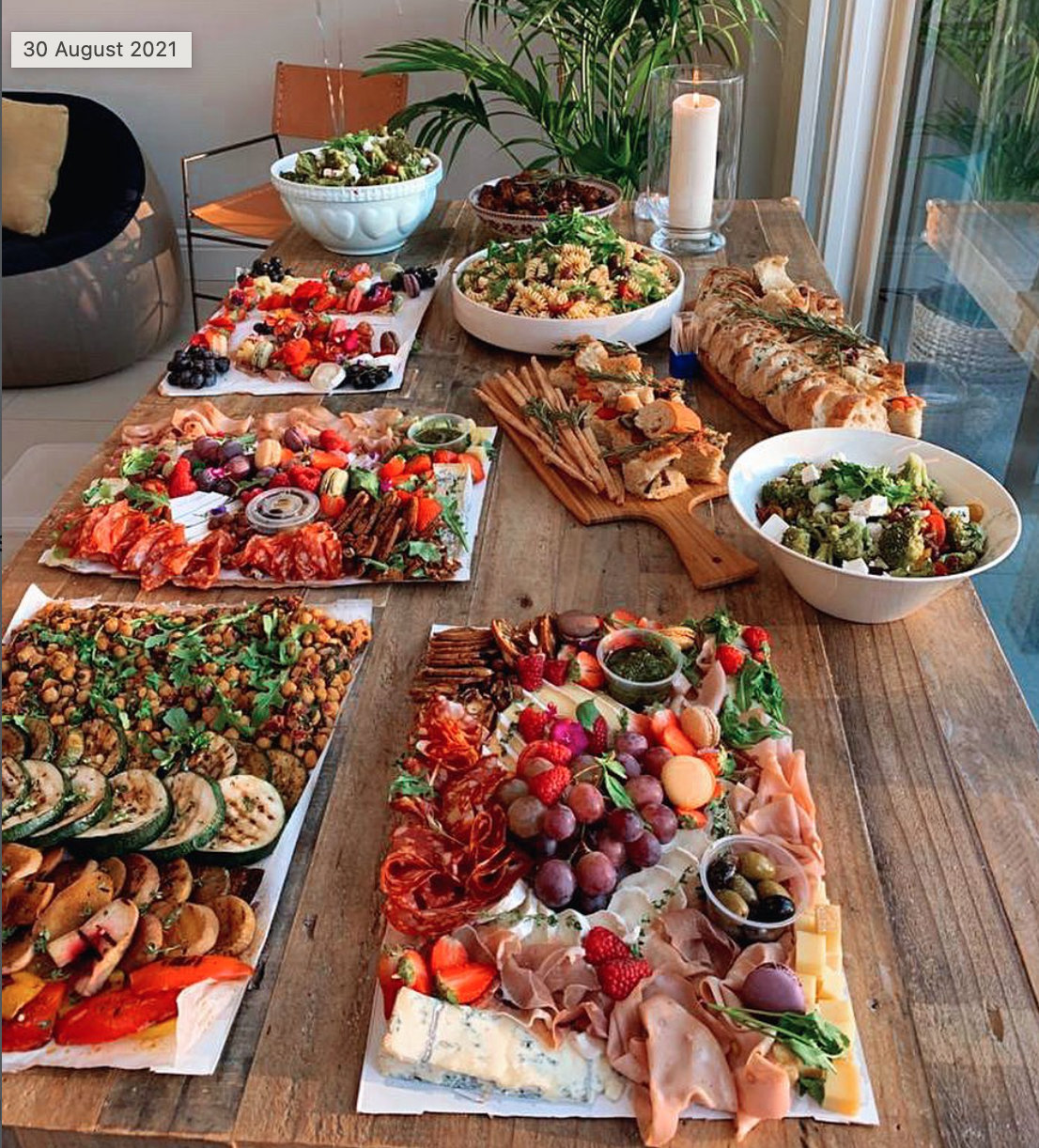 A wooden table filled with various platters of appetizers, salads, and desserts, set in a bright room with a large window and a candle.