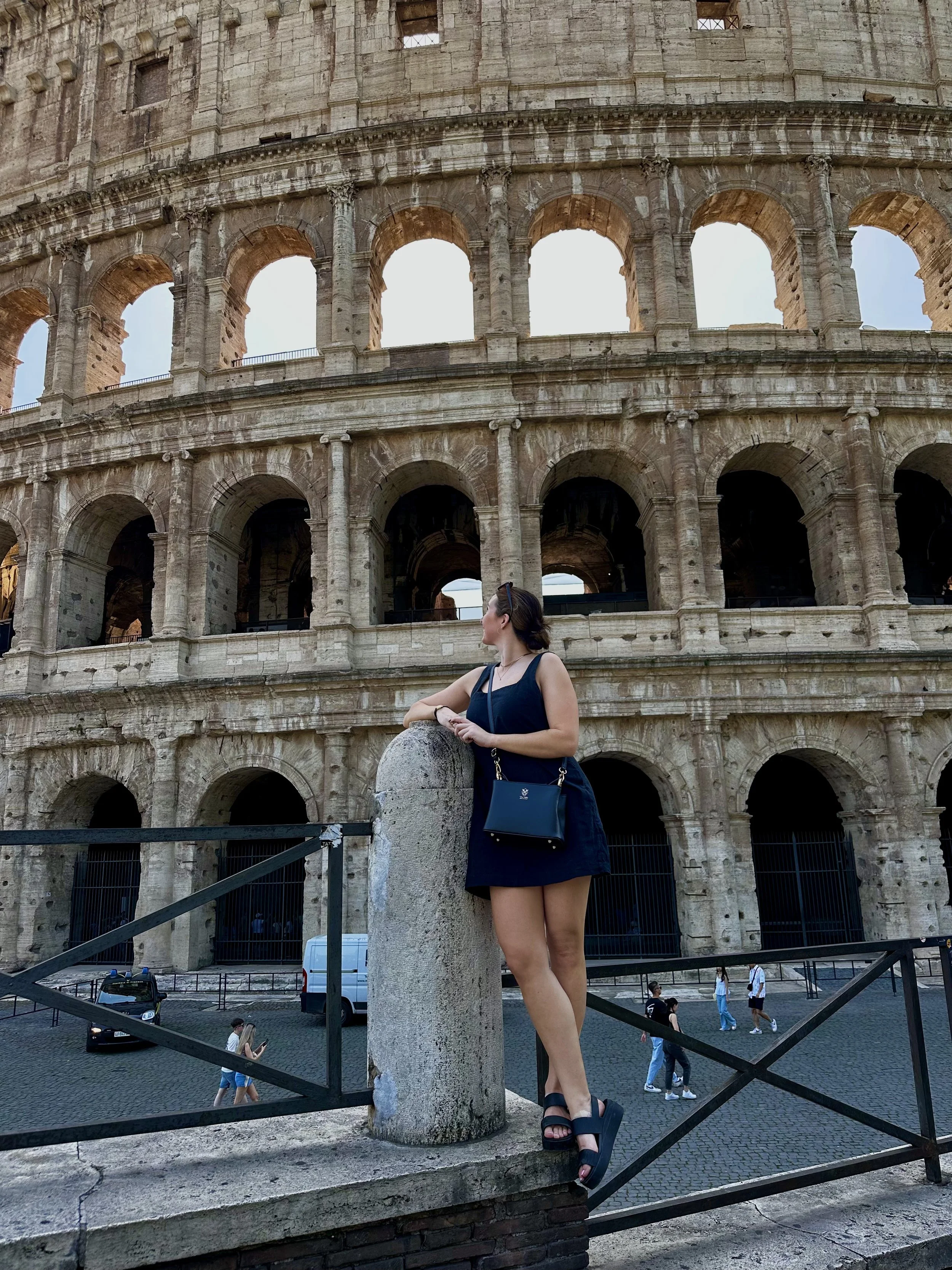 A woman in a black dress and platform sandals stands on a stone ledge, looking to the right, with the Colosseum in Rome in the background.