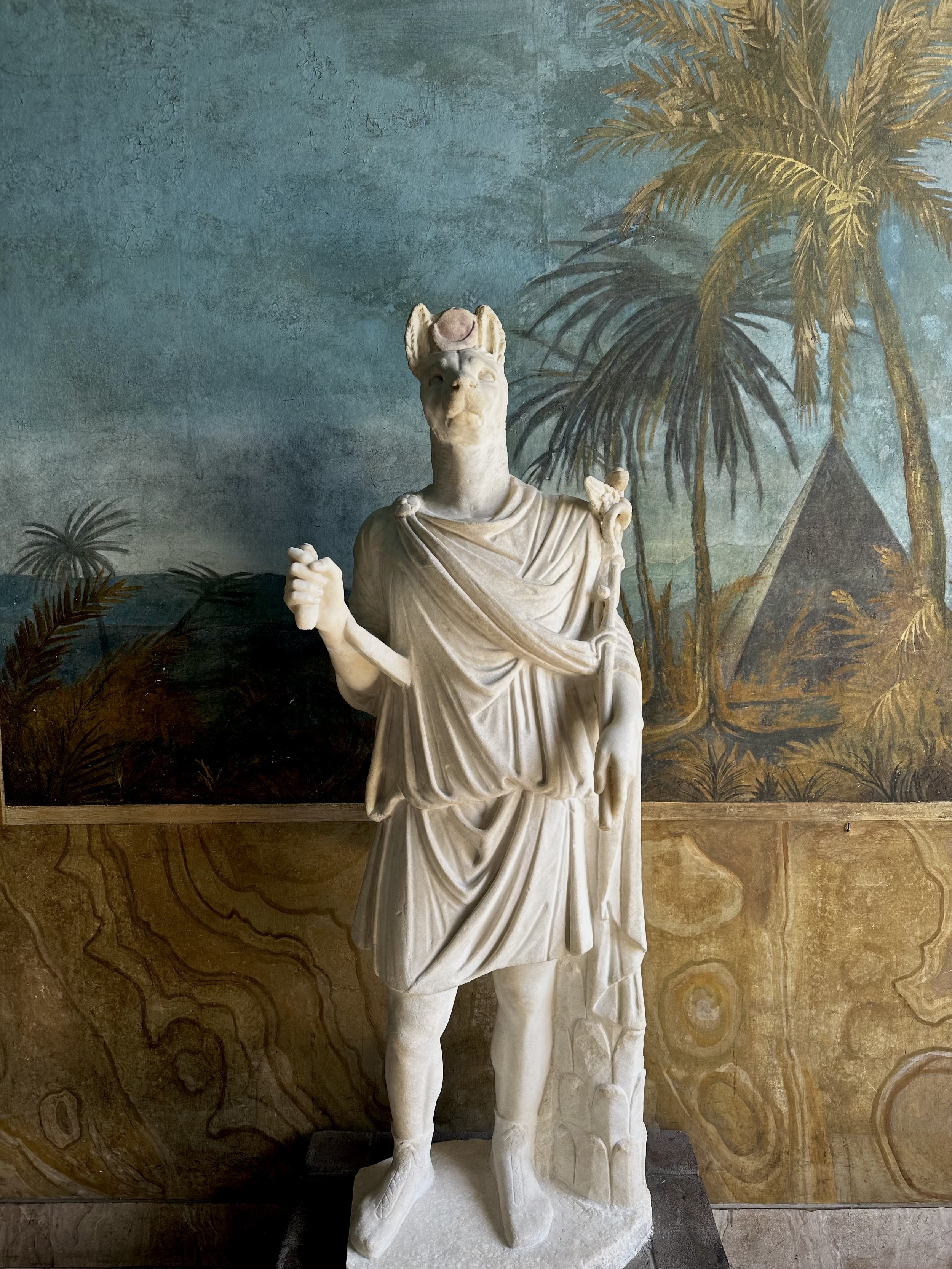A marble statue of a Roman emperor with a dog face wearing a toga, standing outdoors with a painted backdrop of palm trees and a pyramid.