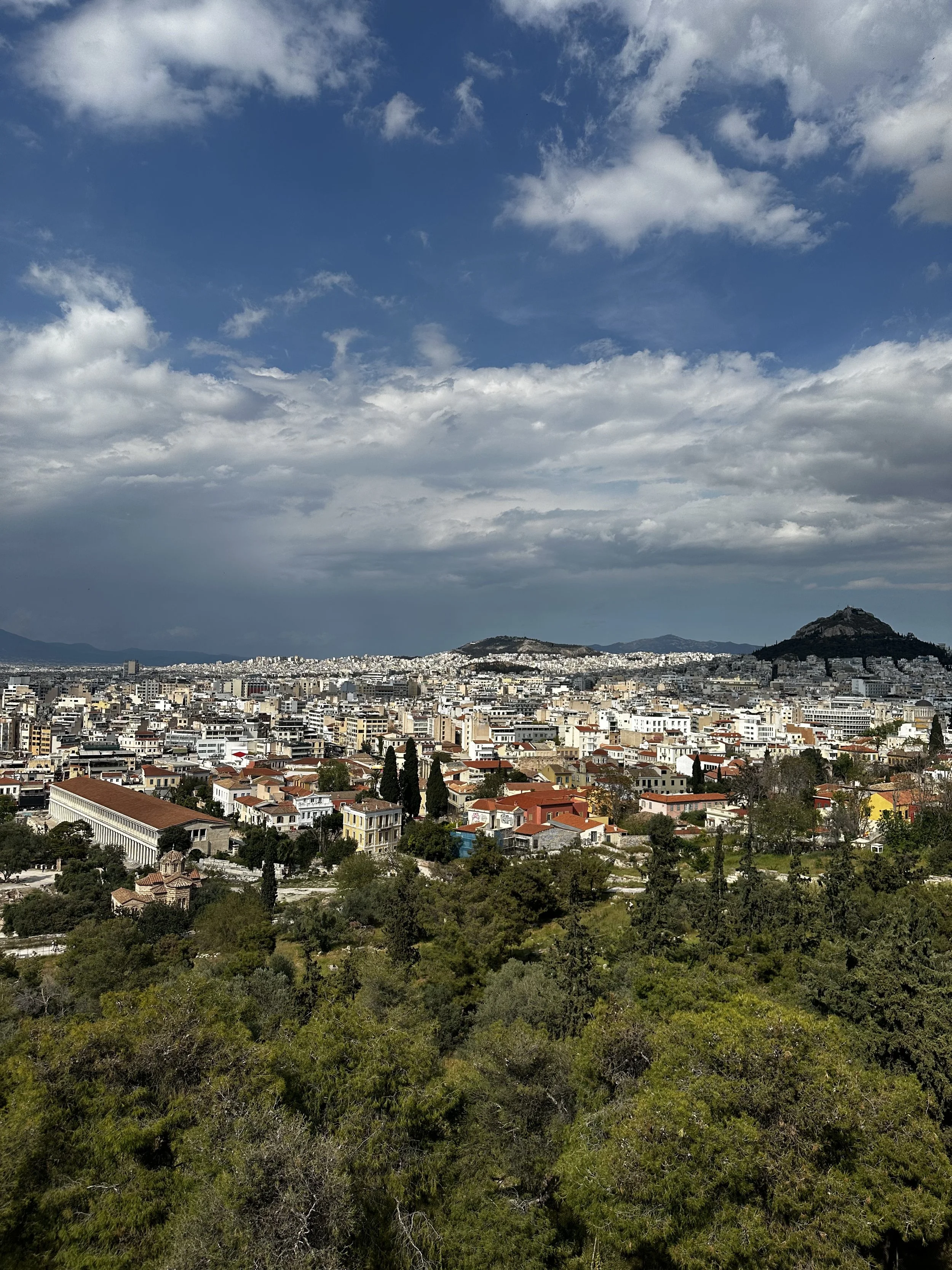 A cityscape under a partly cloudy sky, with green trees in the foreground and numerous buildings extending to the horizon, featuring two prominent hills in the background.