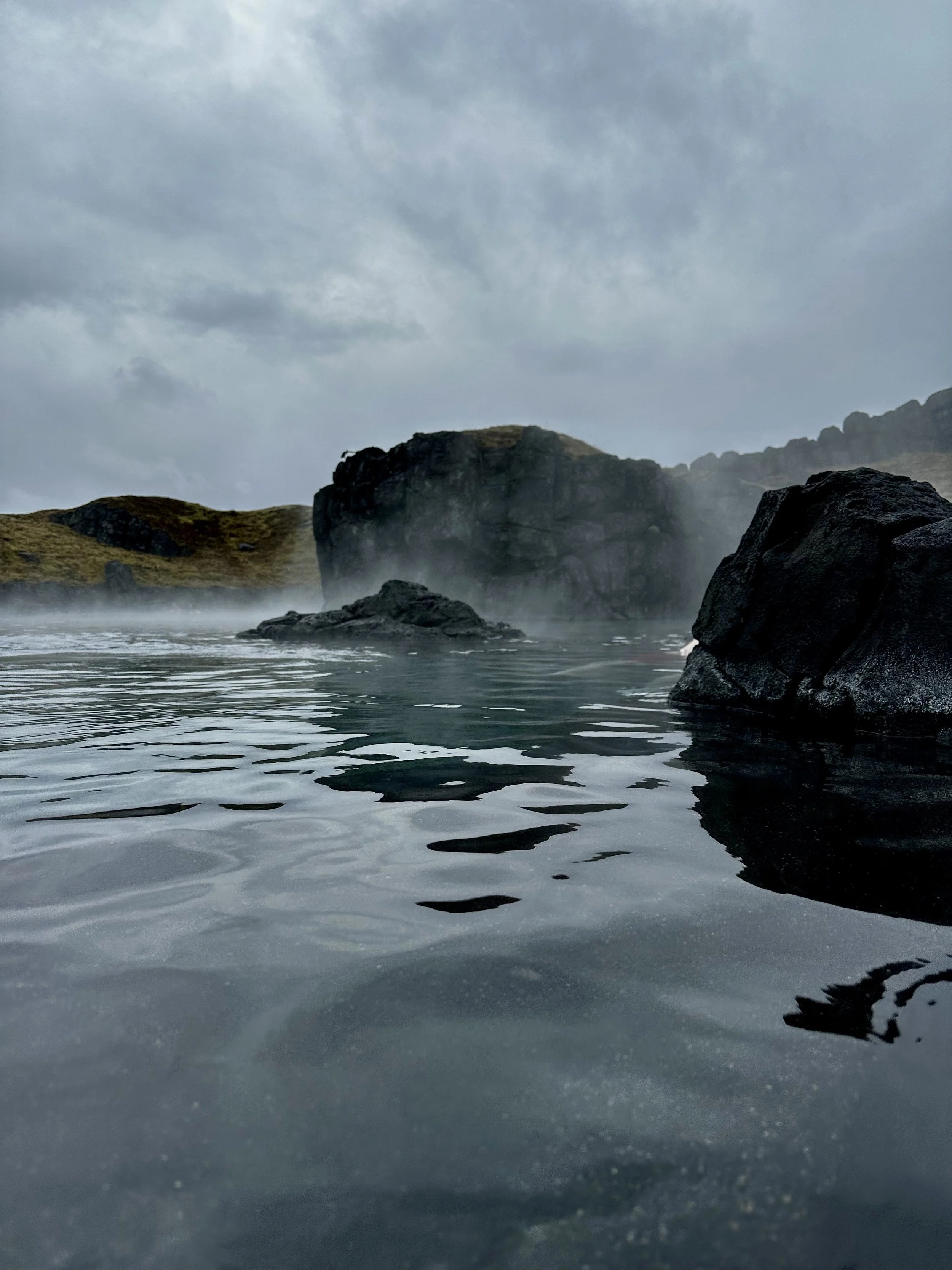 Steam rising from a geothermal hot spring in Iceland, surrounded by dark volcanic rock.