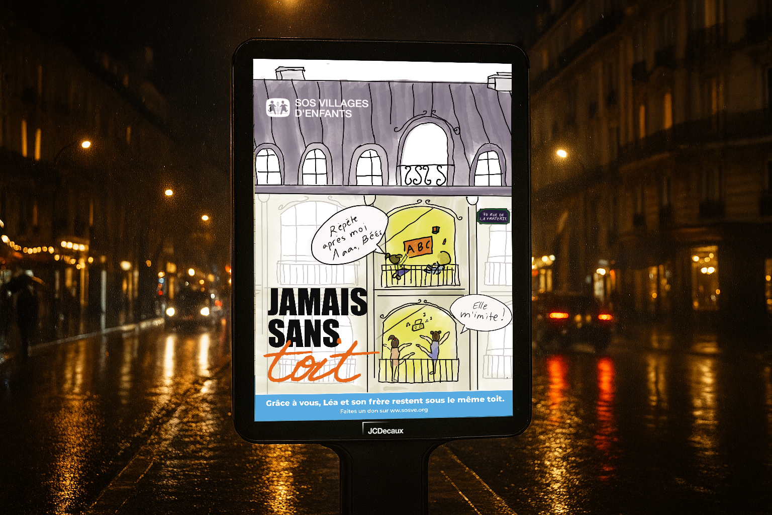 A digital display board showing an illustration of a building with two balconies and kids playing, with text in French and French phrases, and the phrase "JAMAIS SANS tout" in bold black and orange letters, set against a rainy city street at night with reflections of streetlights on the wet pavement.