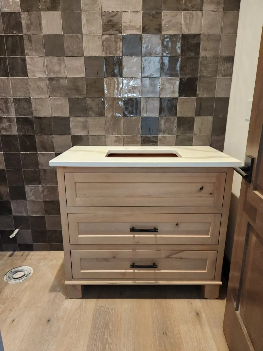A wooden vanity with three drawers and a rectangular cutout on the top for a sink, positioned in front of a tiled wall in a bathroom.