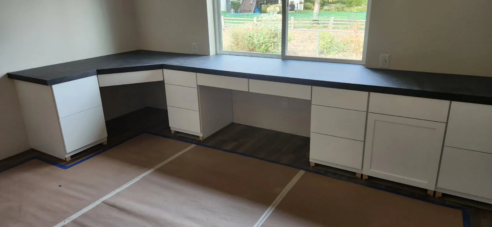 Kitchen with white cabinets and a black countertop, large window overlooking a backyard, and a taped paper floor.