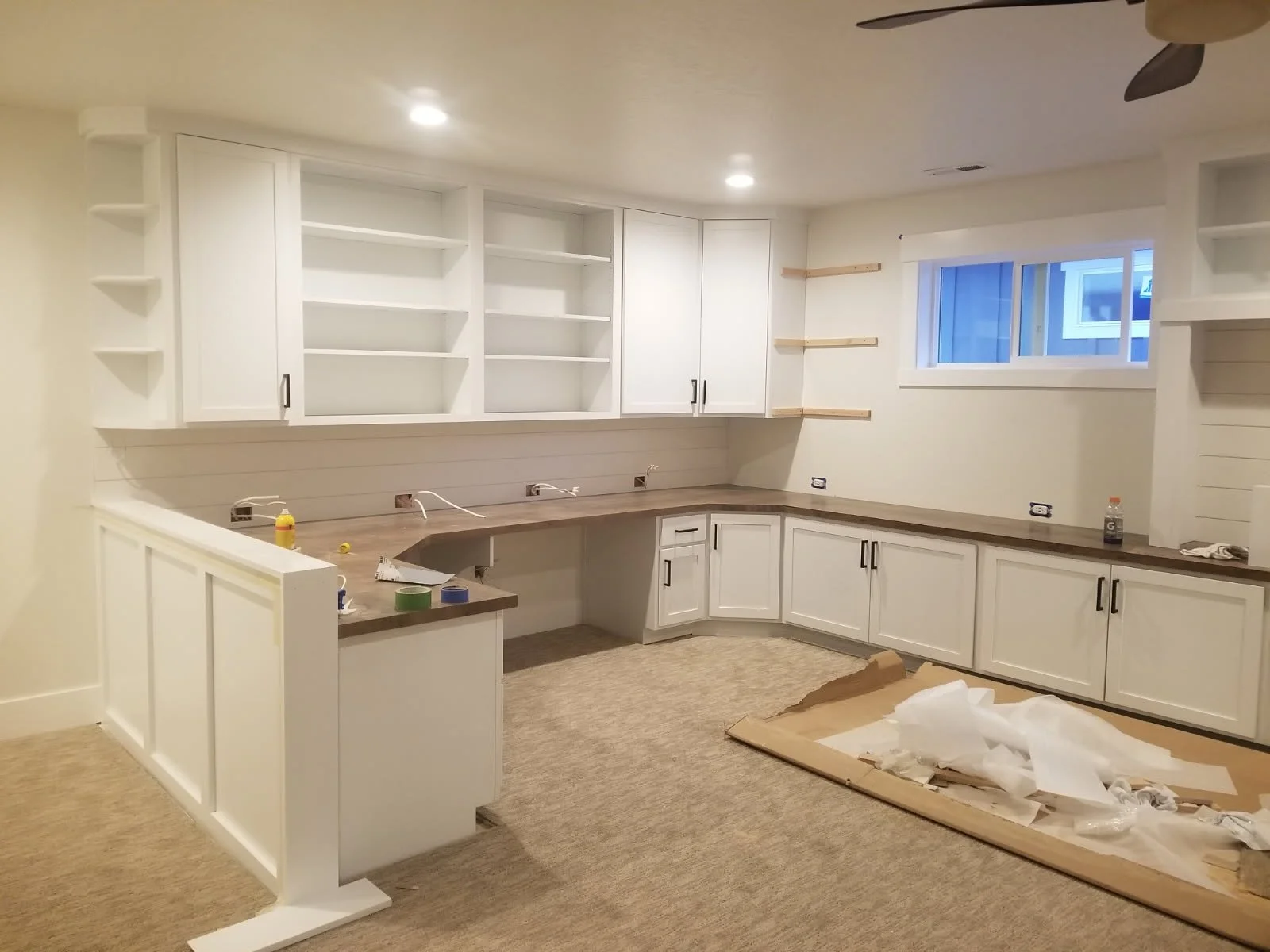 Kitchen under renovation with white cabinets, open shelves, and a window. Items and tools are on the countertop, and a protective sheet with paper debris is on the floor.