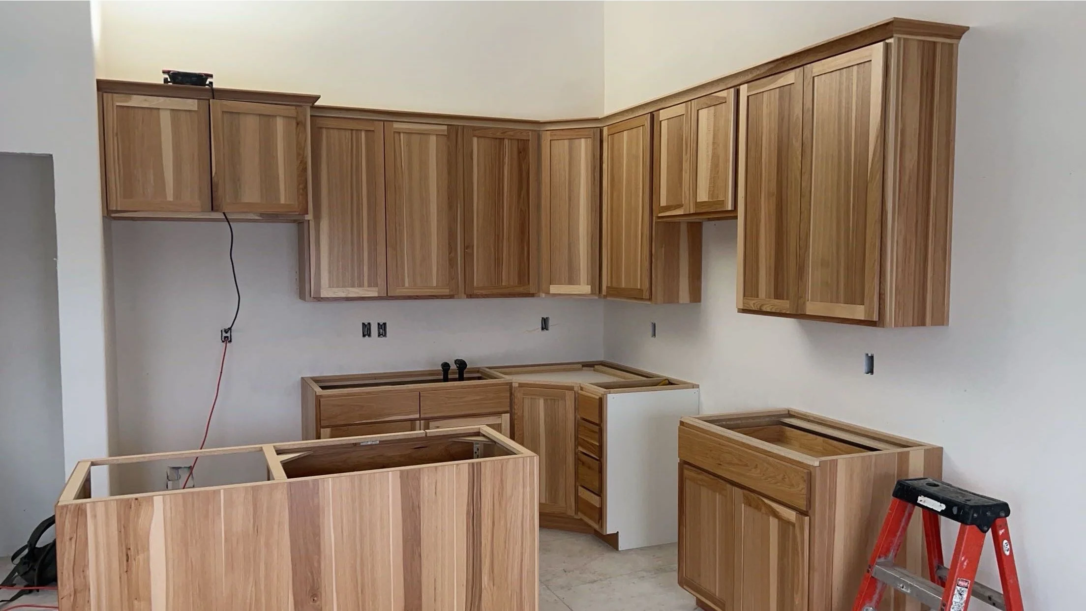Rustic New Kitchen cabinet install with wooden cabinets, some wall cabinets installed, and unfinished lower cabinets. A red step ladder is present, and construction tools and wires are visible.