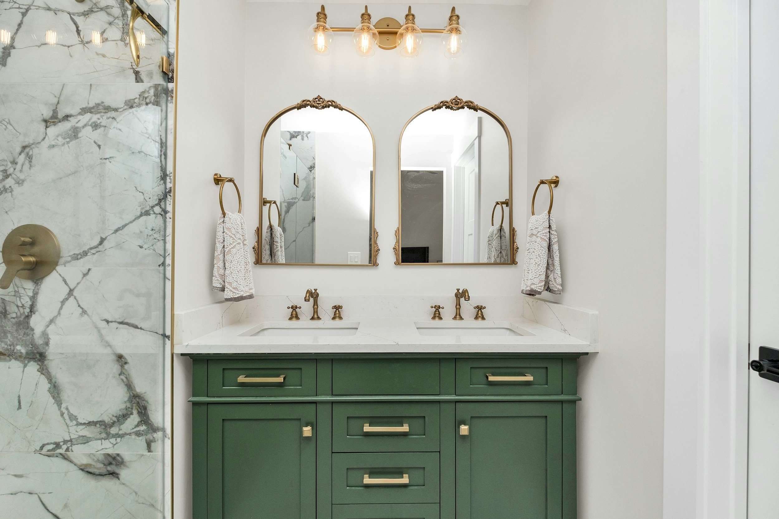 Double vanity bathroom with green cabinets, white marble countertop, two brass faucets, two ornate brass-framed mirrors, wall-mounted brass towel rings with white towels, marble shower on the left, four-bulb brass light fixture above the mirrors.