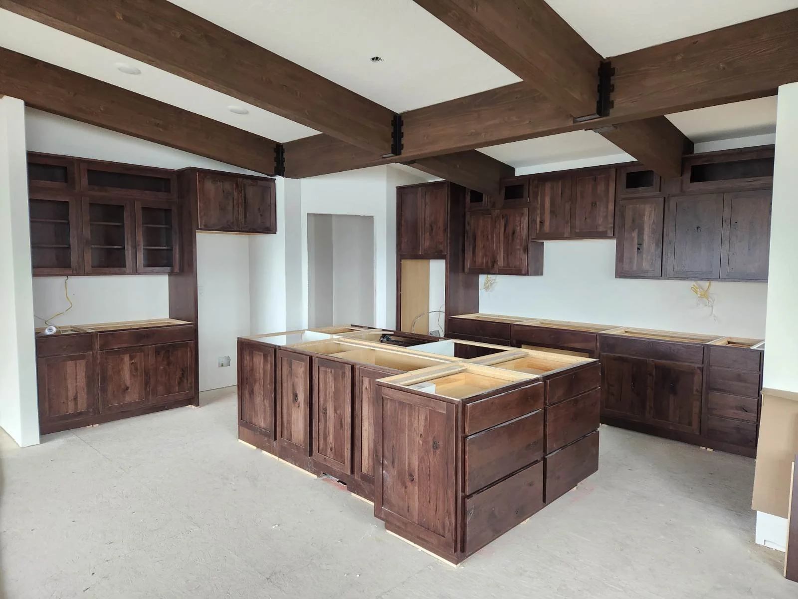 Kitchen cabinets and island under construction, with dark wood finish, open spaces for countertops, and a ceiling with exposed wood beams.