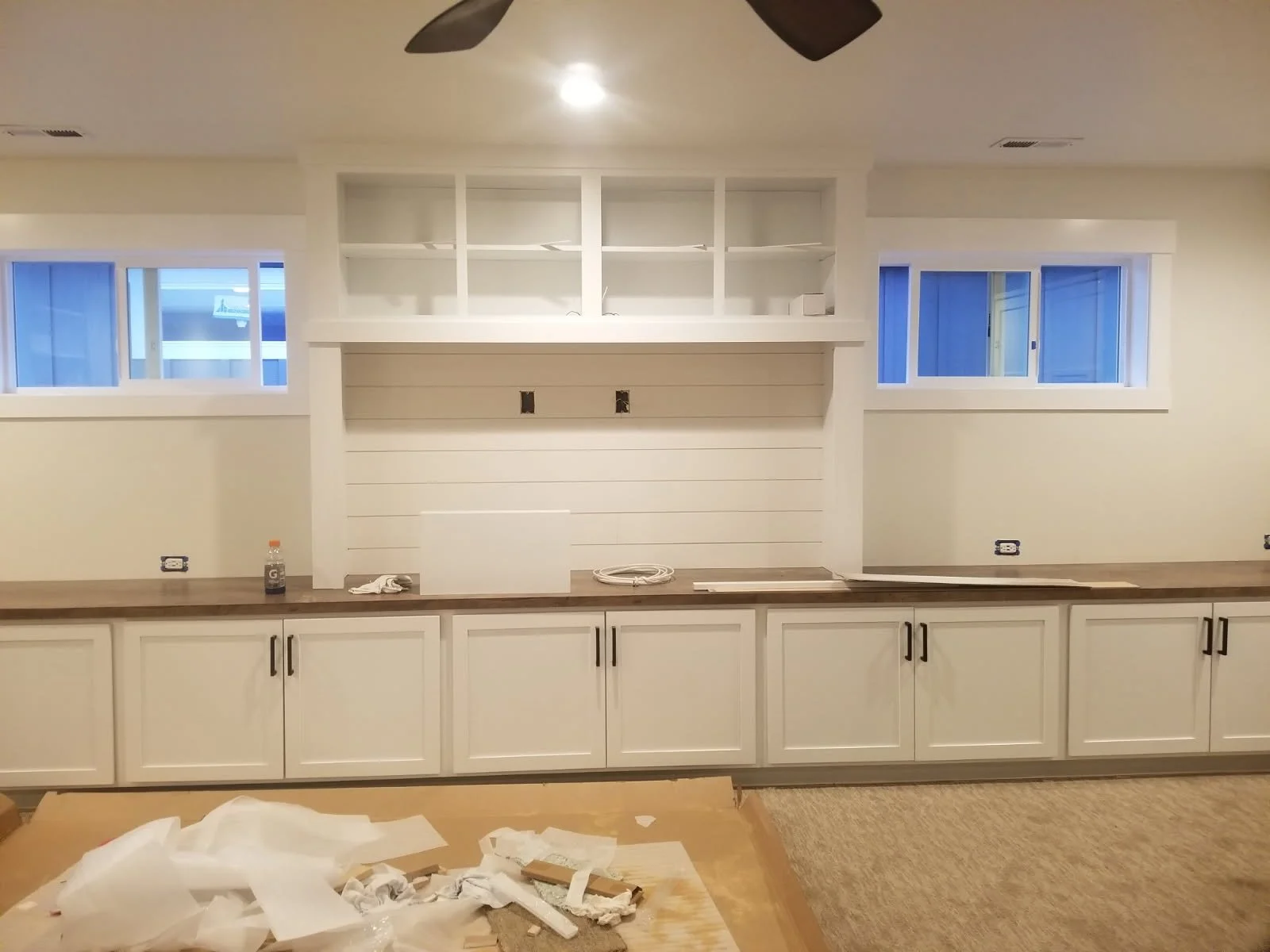 Kitchen with white cabinets, wooden countertop, and two top windows, with construction materials and clutter on the floor.